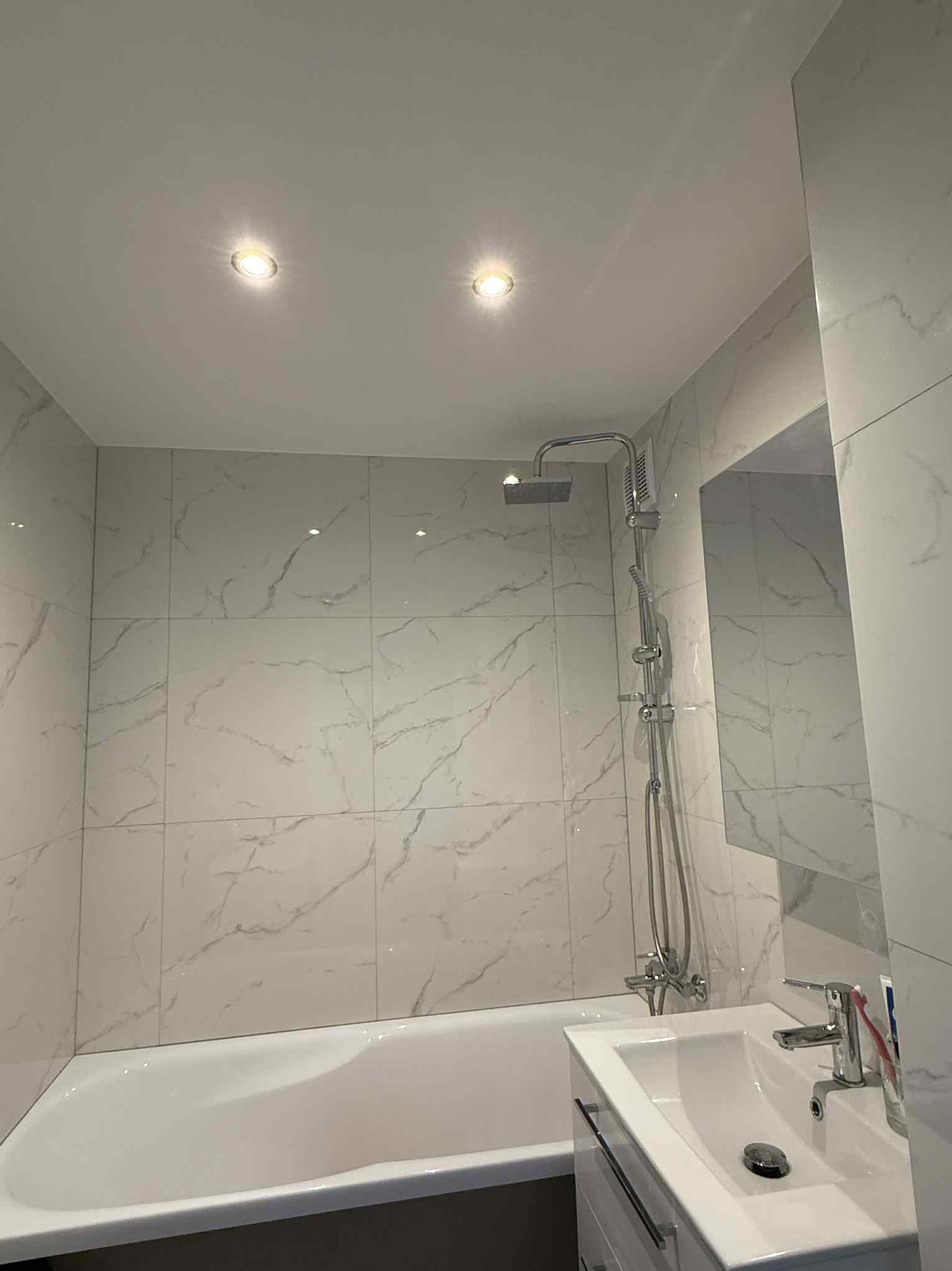 Wykończona, jasna łazienka z wanną, umywalką i marmurowymi płytkami. Widoczne oświetlenie punktowe i chromowana armatura. Minimalistyczny design.