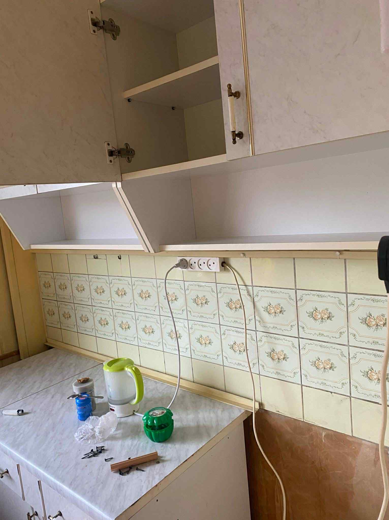 Kuchenne szafki z otwartymi drzwiczkami i beżowymi płytkami z motywem kwiatowym. Na blacie czajnik, pojemniki i drobne narzędzia.