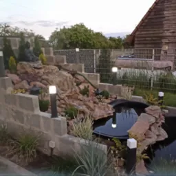 Oświetlony ogród z kaskadą wodną i stawem. Lampy ogrodowe wzdłuż ścieżki i na murku. W tle drewniany budynek i zielona roślinność. Kamienna dekoracja.