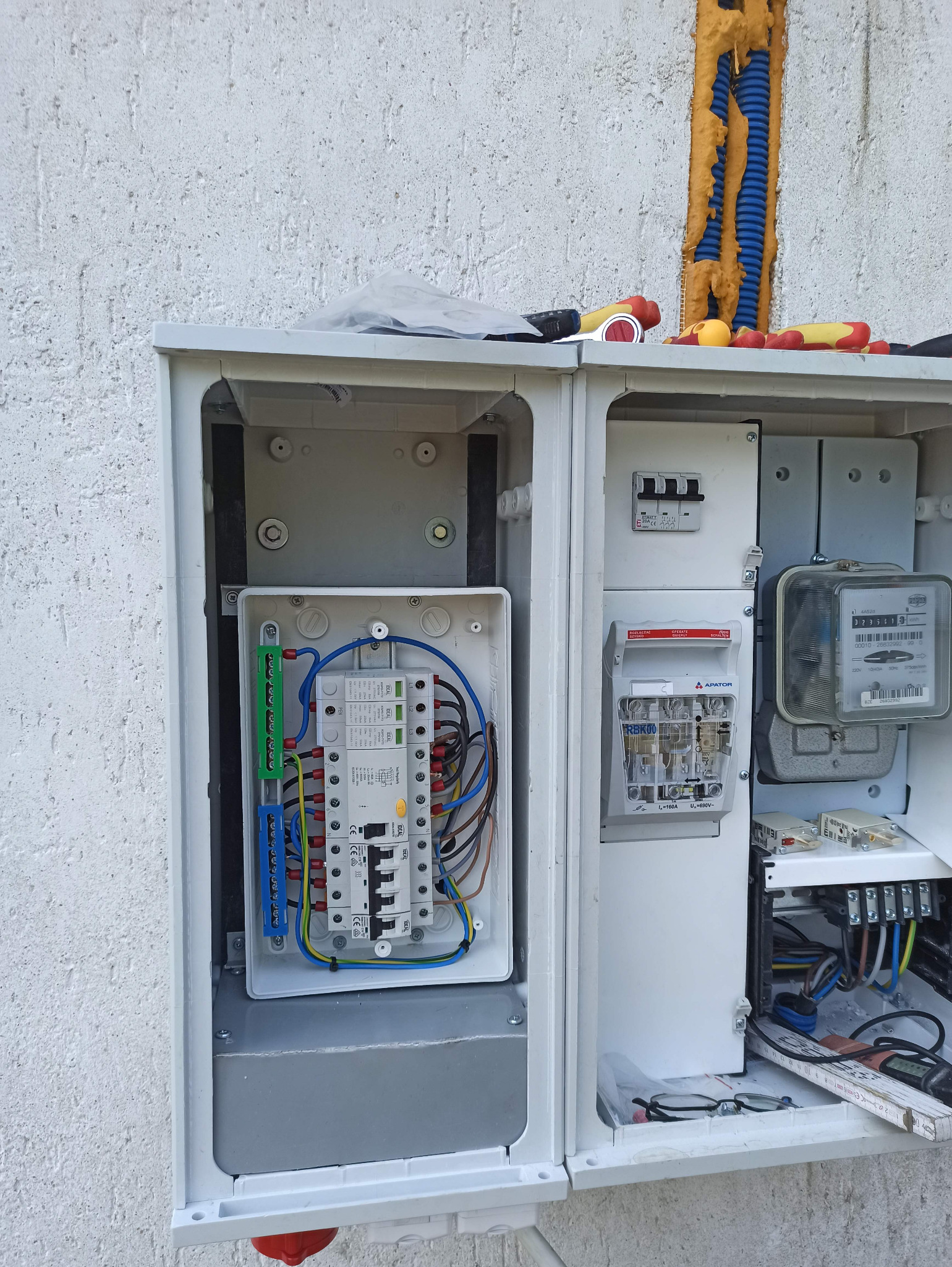 Otwarta, biała rozdzielnica zewnętrzna z układem kontrolno-pomiarowym i zabezpieczeniami, zamontowana na szarej ścianie budynku. Widoczne przewody, bezpieczniki i licznik energii.