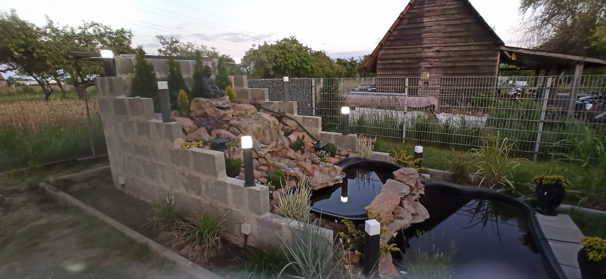 Oświetlony ogród z kaskadą wodną i stawem. Lampy ogrodowe wzdłuż ścieżki i na murku. W tle drewniany budynek i zielona roślinność. Kamienna dekoracja.