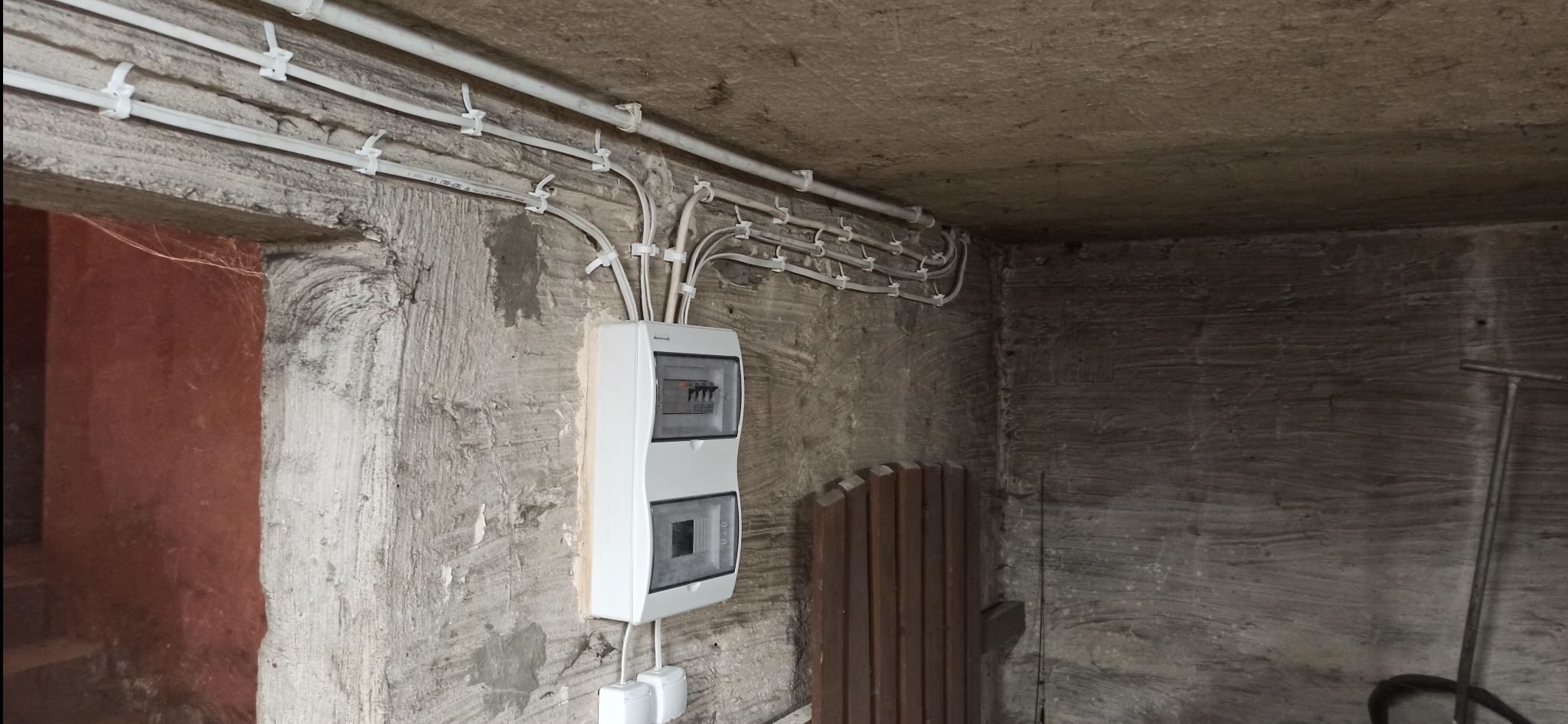 Natynkowa rozdzielnica z przewodami w białych rurkach na szarej, chropowatej ścianie piwnicy. Widoczne gniazda i drewniane elementy.