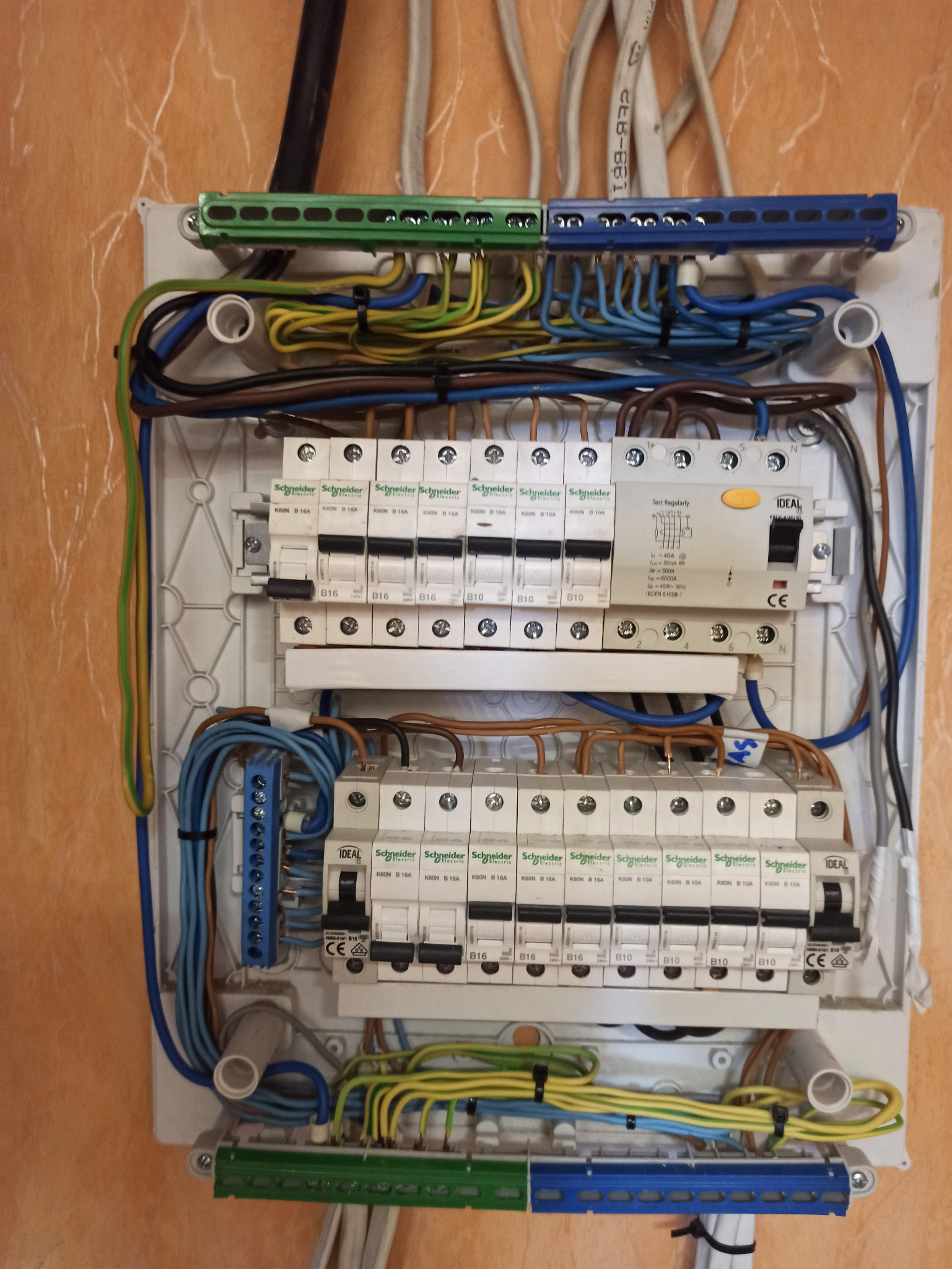 Rozdzielnica elektryczna z widocznymi bezpiecznikami, przewodami i listwami zaciskowymi, zamontowana na ścianie o jasnej strukturze. Widok z bliska, detale instalacji.