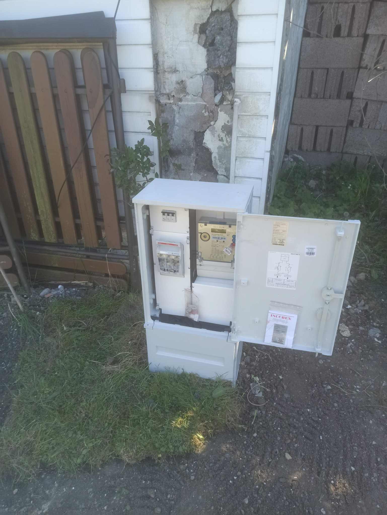 Otwarta skrzynka elektryczna z licznikiem na trawie, obok ściany z uszkodzonym tynkiem. Widoczne elementy instalacji wewnątrz szafki, schemat na drzwiczkach.