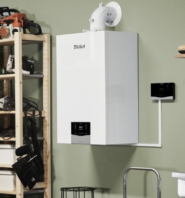 Biały piec gazowy Vaillant wiszący na ścianie, z podłączonym sterownikiem, na tle półki z narzędziami i drabiny. Widoczny komin.