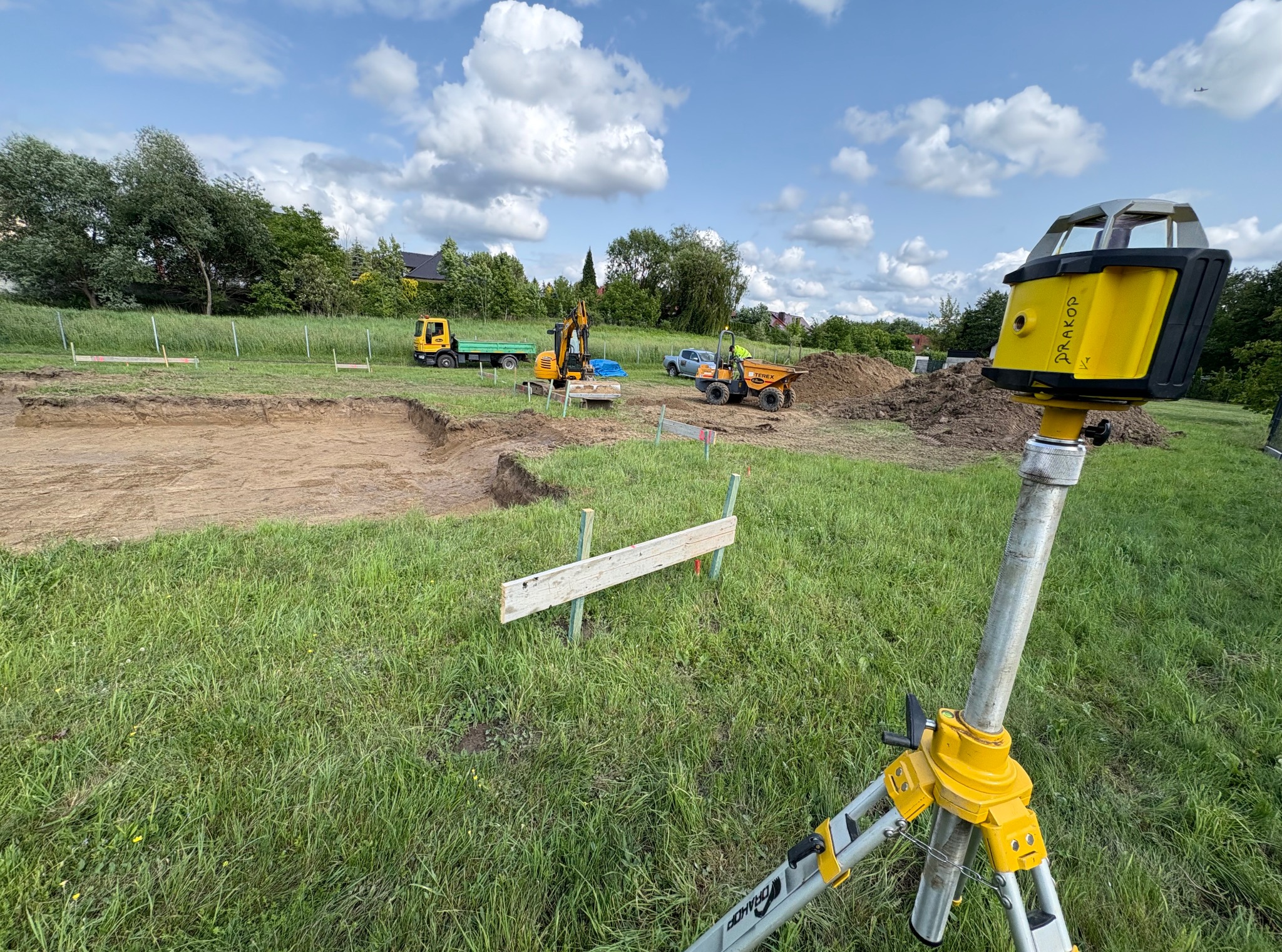 Wykonany wykop pod fundamenty z użyciem niwelatora laserowego, w tle maszyny budowlane i wywrotka. Teren zielony, częściowo przygotowany pod budowę.