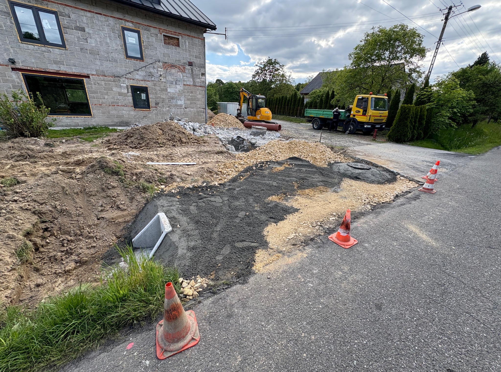 Prace ziemne przy domu z użyciem koparki i wywrotki. Widoczne ułożone rury, gruz i stożki ostrzegawcze. Etap przygotowania terenu pod dalsze prace budowlane.