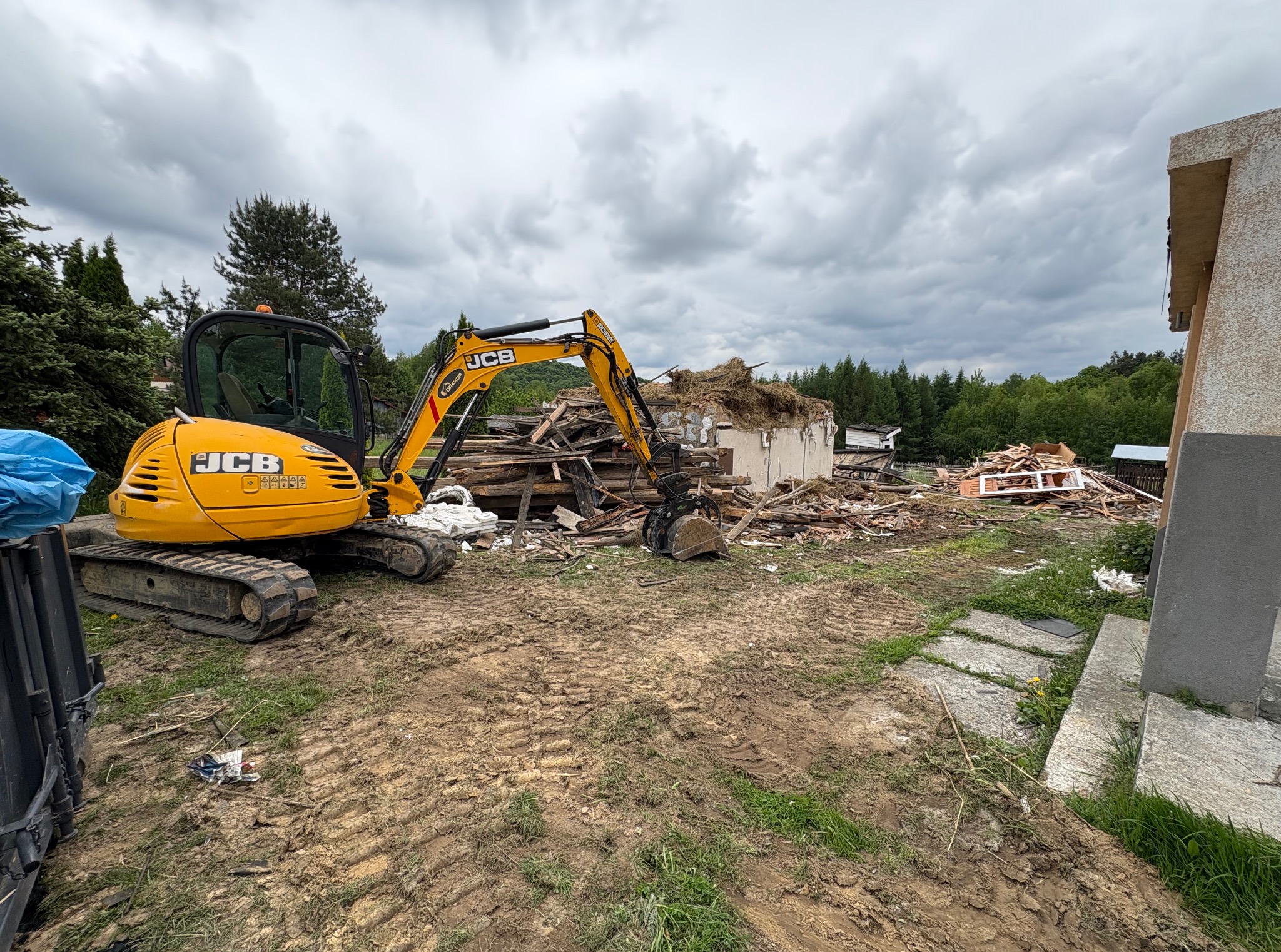 Żółta koparka JCB przy rozbiórce budynku w Niepołomicach. Widoczne gruzy i fragmenty konstrukcji. Niebo pochmurne.