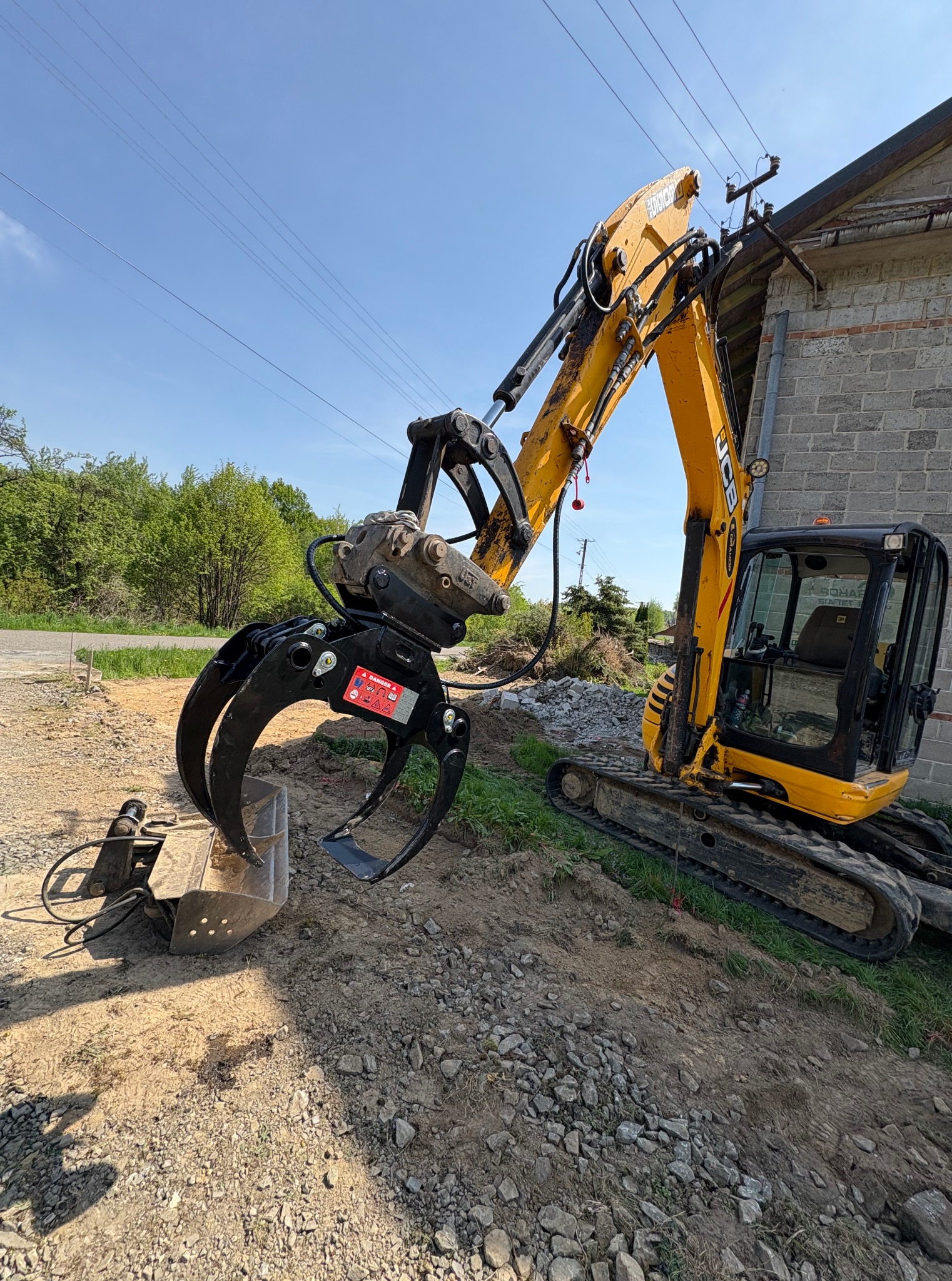 Żółta koparka JCB z hydraulicznymi chwytakami, stojąca na gruzowisku obok murowanego budynku. Widoczny fragment drogi i zieleń w tle, błękitne niebo.
