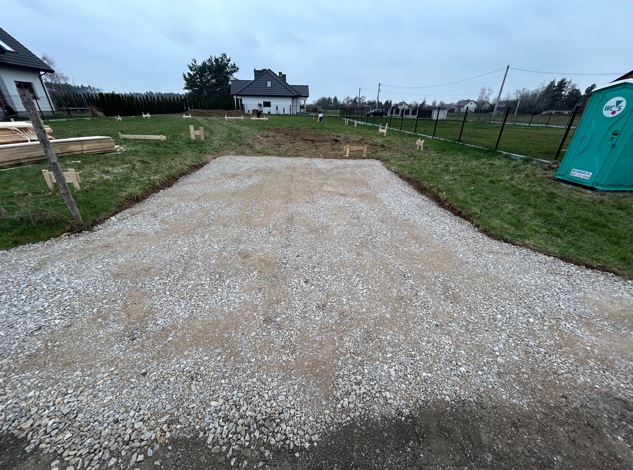 Utrwardzony teren z kruszywa na działce, przygotowany pod budowę. W tle widoczny dom jednorodzinny i przenośna toaleta.