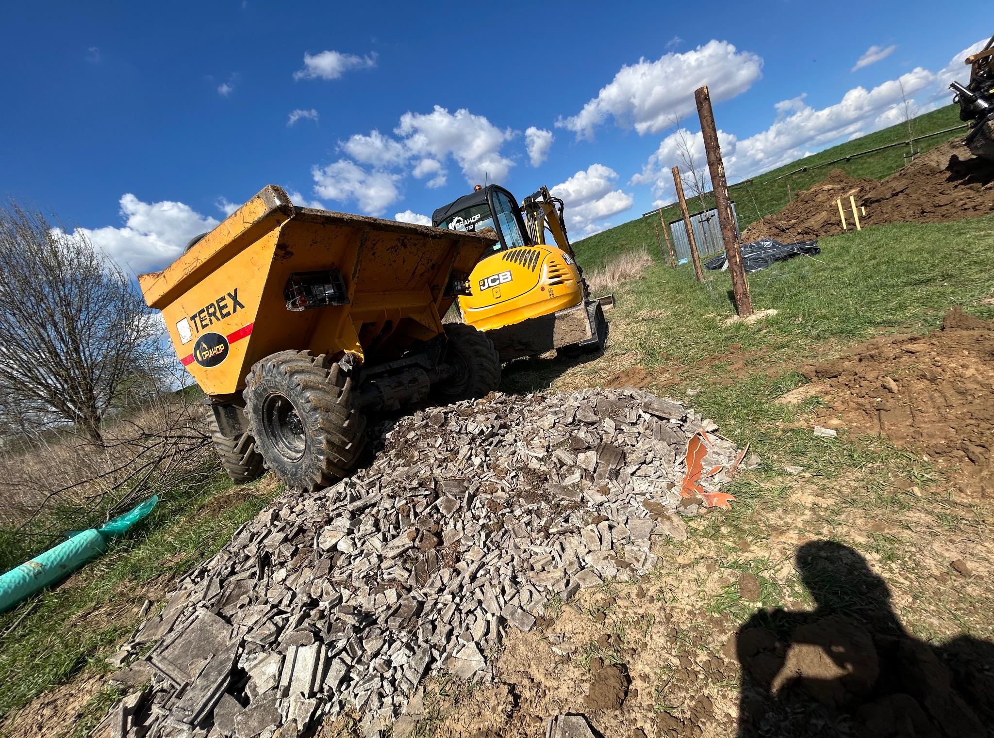 Żółta wywrotka Terex i koparka JCB na nierównym terenie z gruzem i ziemią, na tle błękitnego nieba z chmurami. Widoczne ślady prac ziemnych i wykopów.