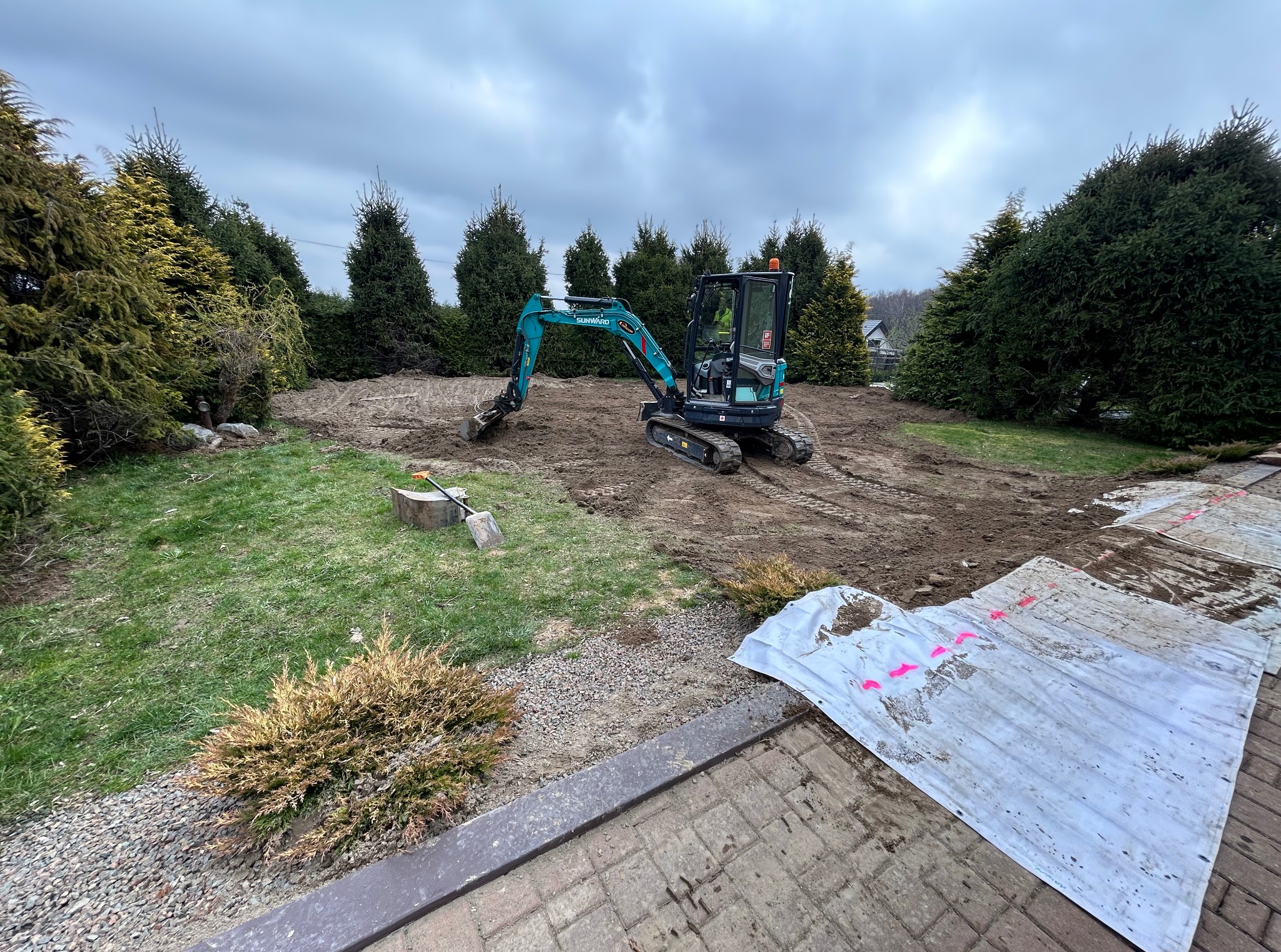 Niebieska mini koparka Sunward na częściowo wyrównanym terenie działki, z widocznymi śladami pracy i zielenią w tle, pod pochmurnym niebem.