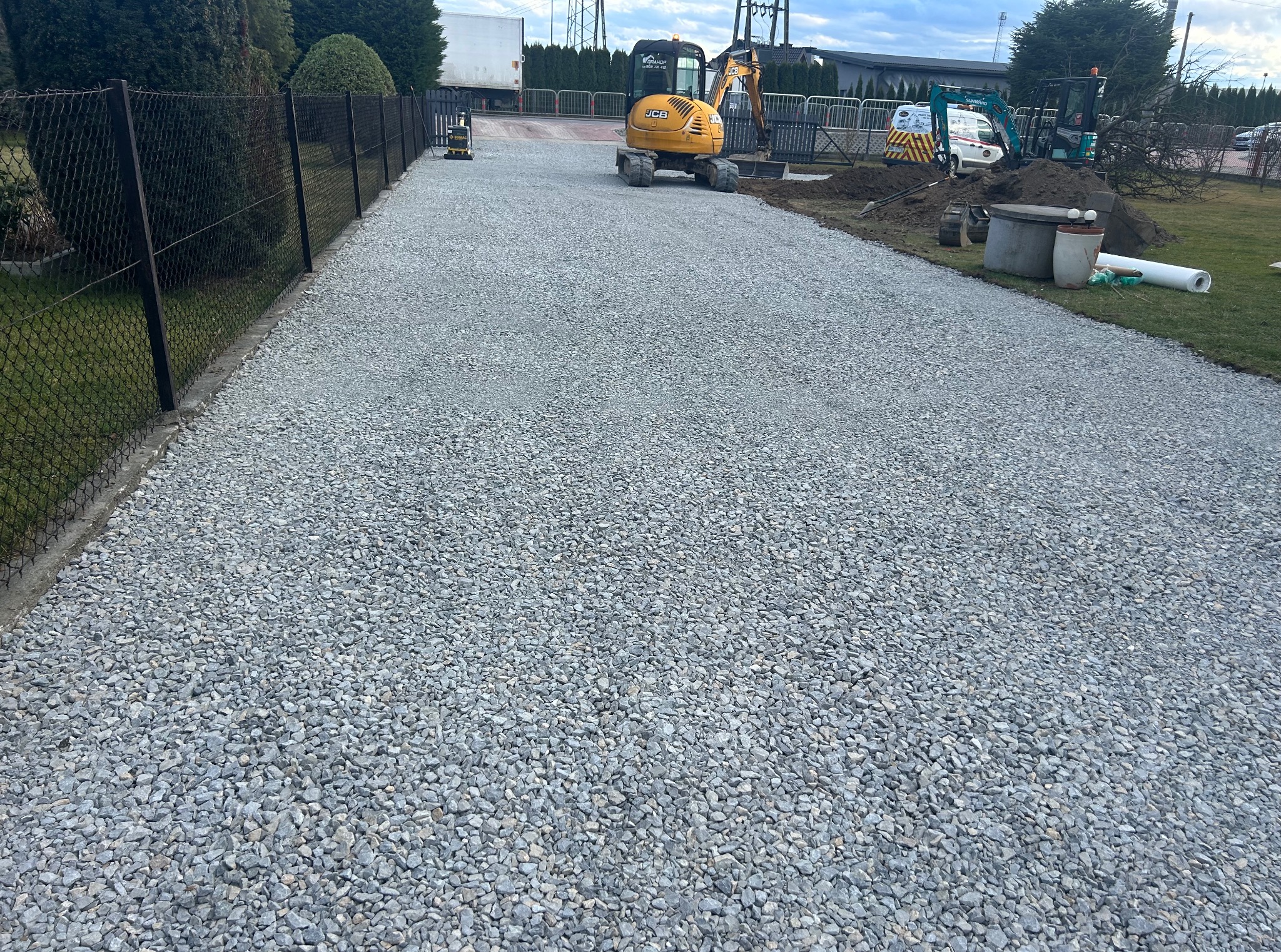 Utwardzona żwirowa droga dojazdowa z żółtą mini koparką JCB w tle. Widoczny fragment czarnego ogrodzenia i zaparkowane pojazdy. Dalsze prace ziemne w tle.