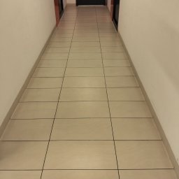 Svitlana Krysa - Wyczyszczony, długi korytarz z beżowymi płytkami podłogowymi i białymi ścianami, prowadzący do czarnych drzwi. Widok z perspektywy podłogi.