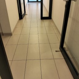 Svitlana Krysa - Korytarz biurowy z beżową podłogą wyłożoną kwadratowymi płytkami. Widoczne otwarte drzwi z czarnymi ramami. Perspektywa z dołu.