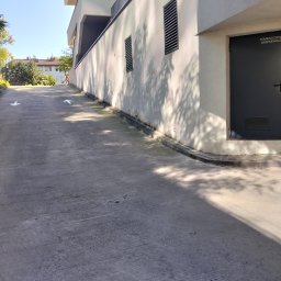 Svitlana Krysa - Zewnętrzna ściana budynku z betonową ścieżką, strzałki kierunkowe na posadzce, cień drzewa na ścianie, drzwi do pomieszczenia gospodarczego.