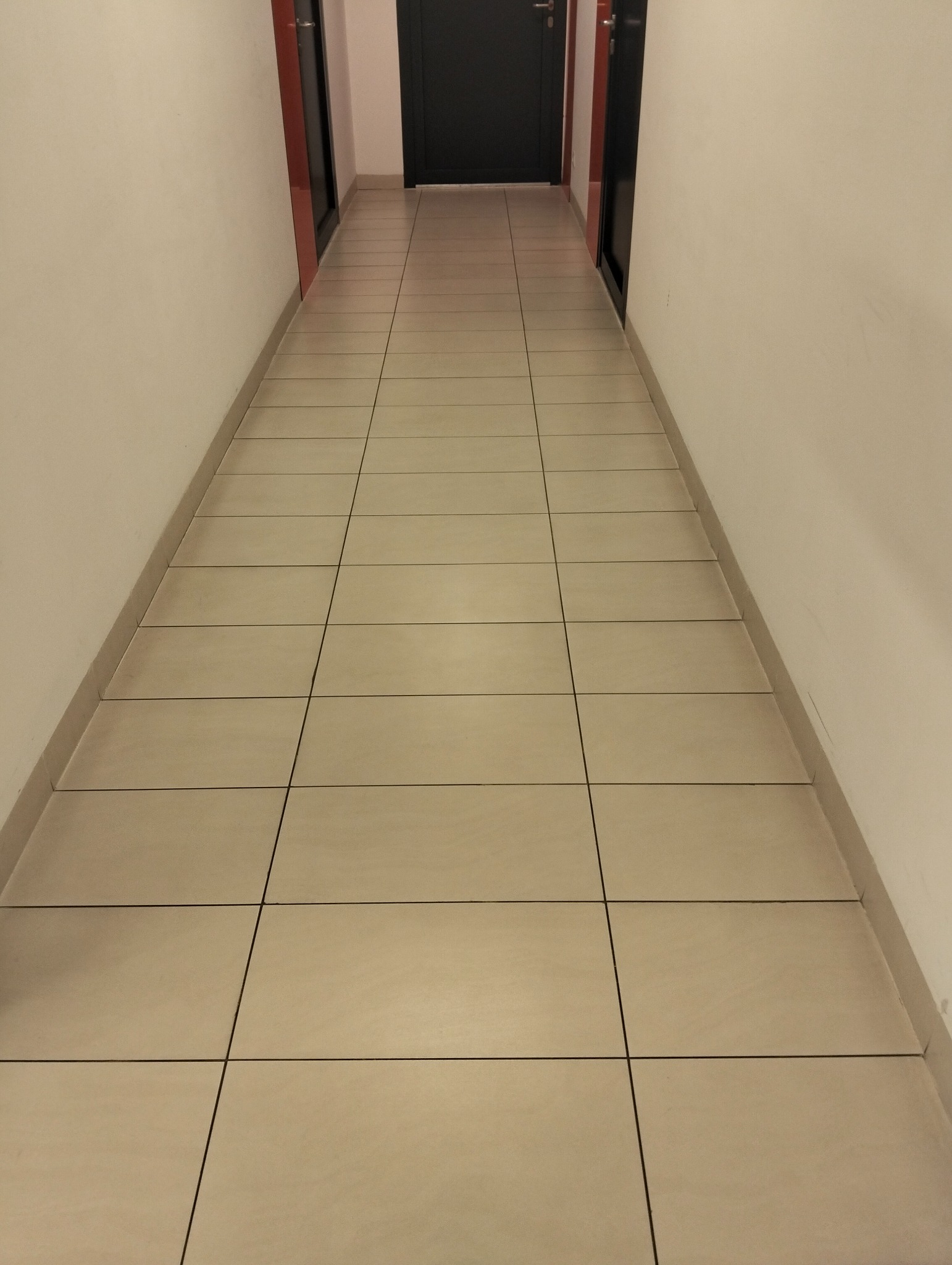 Wyczyszczony, długi korytarz z beżowymi płytkami podłogowymi i białymi ścianami, prowadzący do czarnych drzwi. Widok z perspektywy podłogi.
