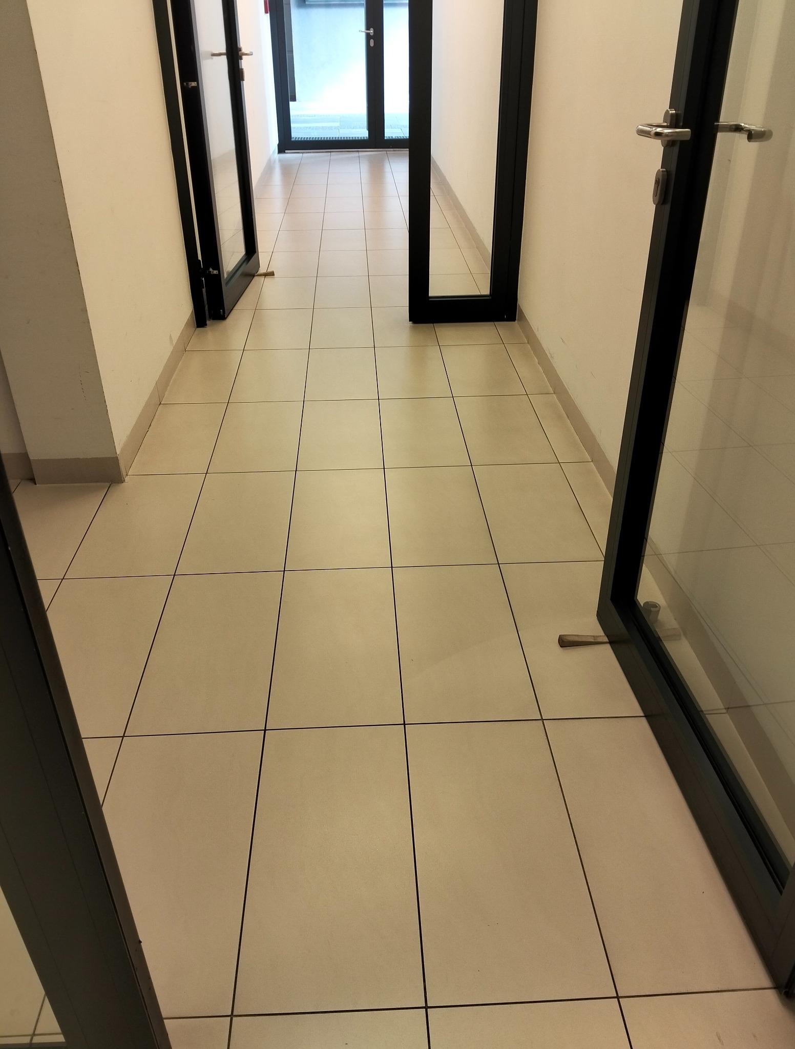 Korytarz biurowy z beżową podłogą wyłożoną kwadratowymi płytkami. Widoczne otwarte drzwi z czarnymi ramami. Perspektywa z dołu.