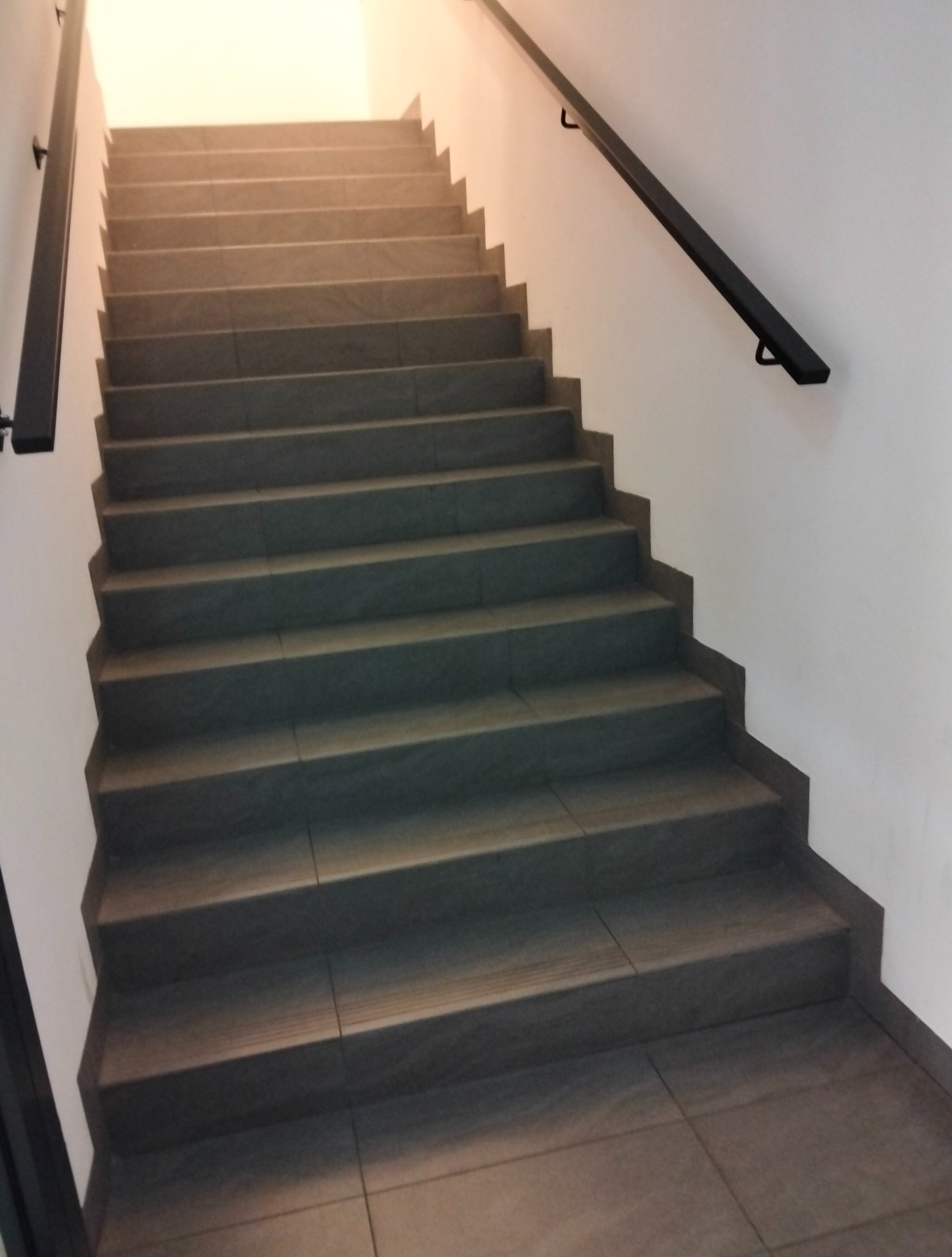 Schody wyłożone szarymi płytkami, z czarną poręczą po prawej stronie, prowadzące do jasnego pomieszczenia. Widok z dołu, lekko z boku.