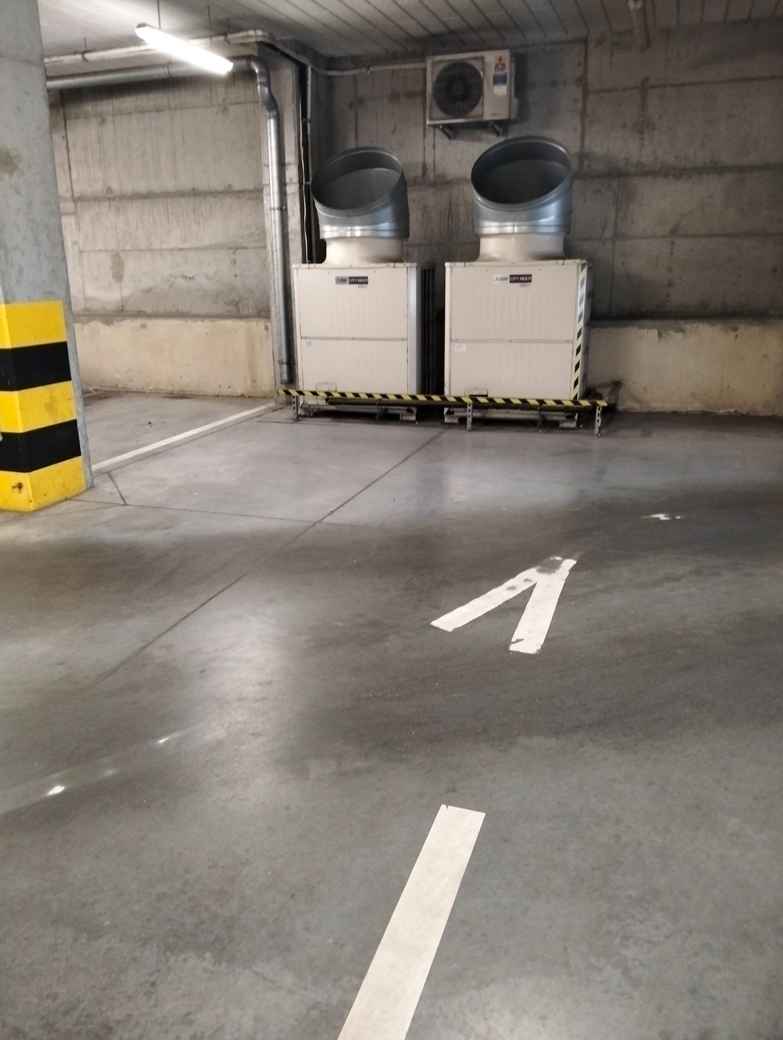 Wnętrze parkingu podziemnego z betonową podłogą, na której widnieją białe strzałki kierunkowe. W tle dwa duże urządzenia klimatyzacyjne.