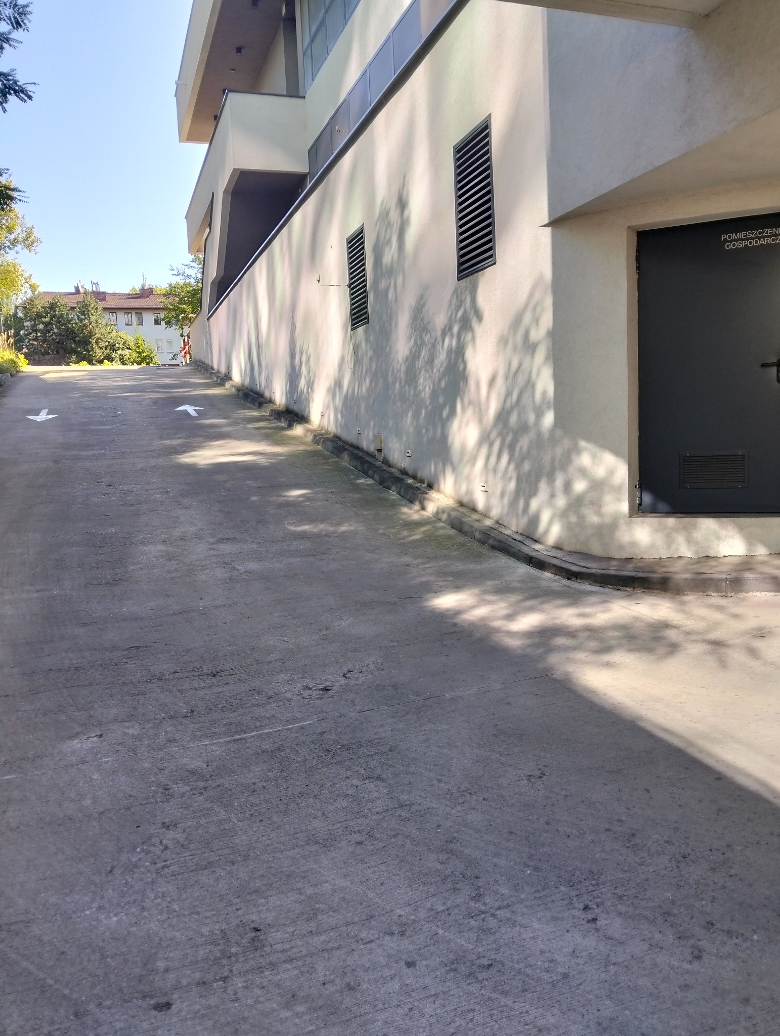 Zewnętrzna ściana budynku z betonową ścieżką, strzałki kierunkowe na posadzce, cień drzewa na ścianie, drzwi do pomieszczenia gospodarczego.