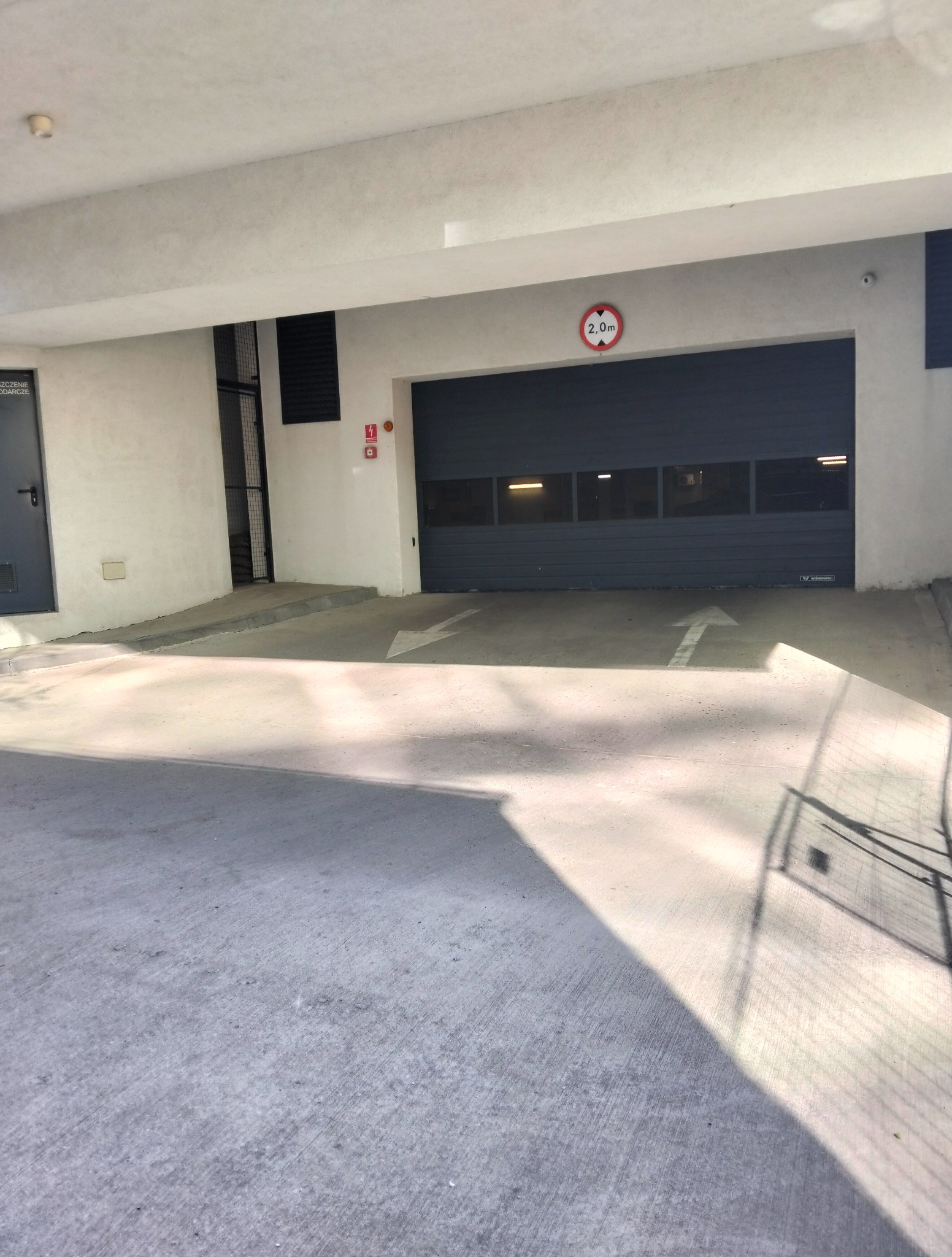 Wjazd do garażu podziemnego z szarą bramą segmentową. Na posadzce betonowej strzałki kierunkowe. Widoczny znak ograniczenia wysokości 2.0m.