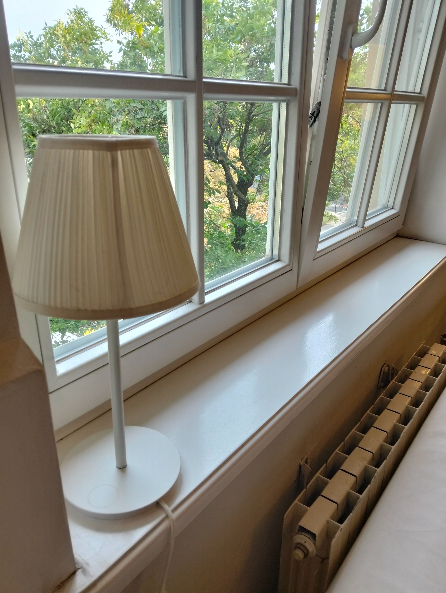 Biały lampion na parapecie okna z widokiem na zieleń. Świeżo pomalowany parapet i grzejnik. Okno z białymi ramami.