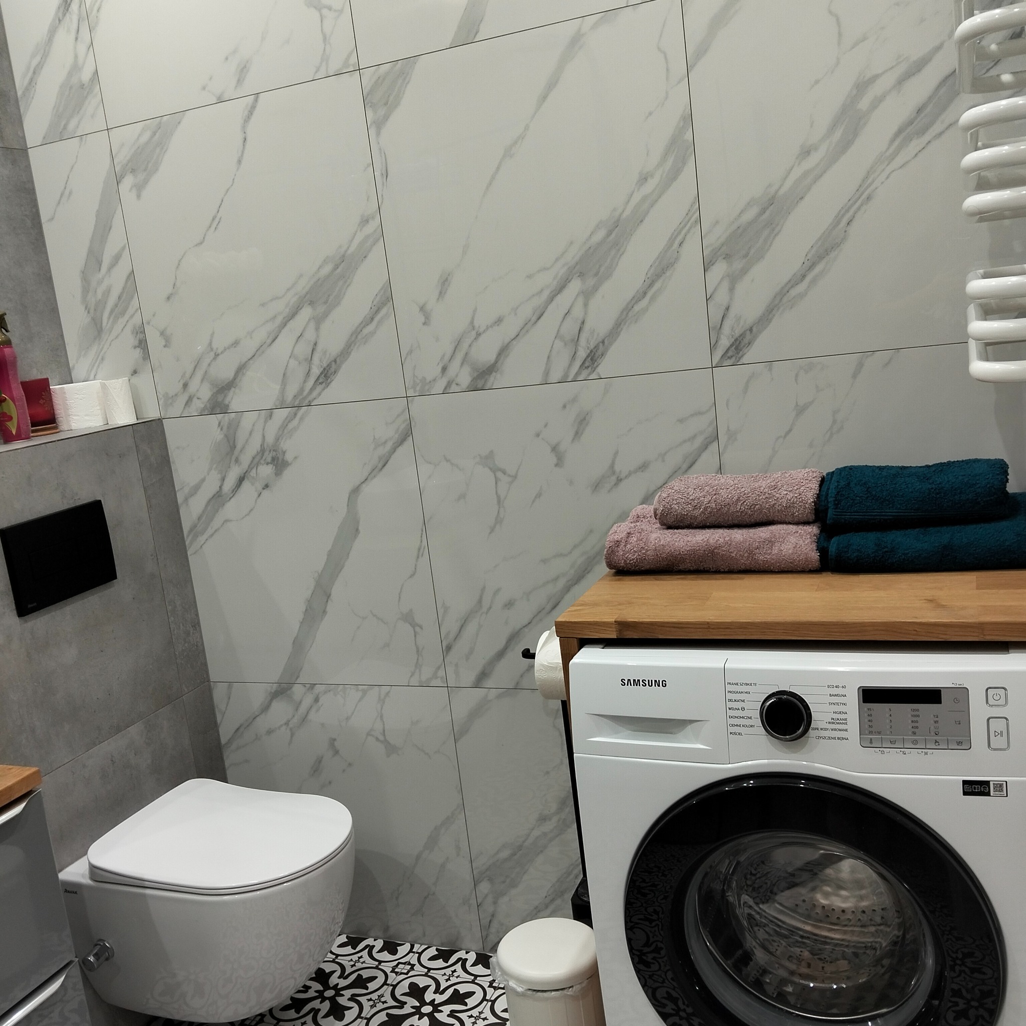 Nowoczesna łazienka z marmurowymi płytkami, wiszącą toaletą i pralką Samsung z blatem, na którym leżą złożone ręczniki. Minimalistyczny design.