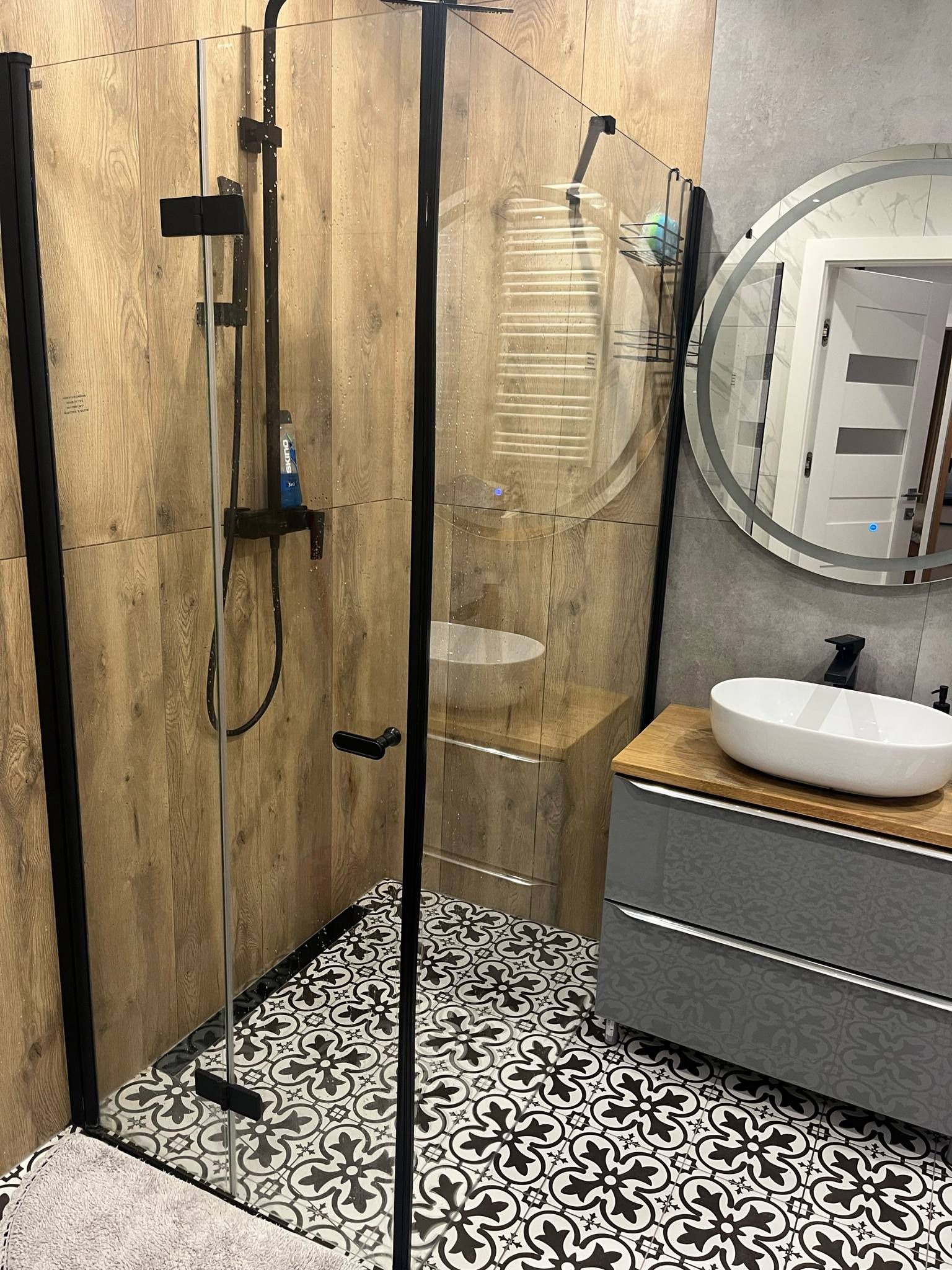 Nowoczesna łazienka z kabiną prysznicową typu walk-in, drewnopodobnymi płytkami ściennymi i wzorzystą podłogą. Umywalka nablatowa i okrągłe lustro z podświetleniem.