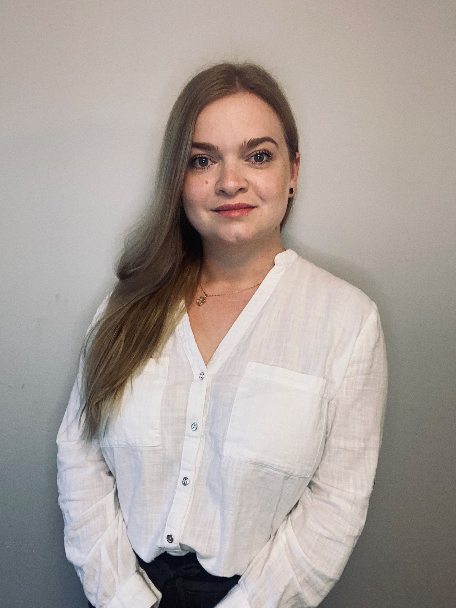 Portret uśmiechniętej kobiety w białej lnianej koszuli z kieszeniami, z delikatnym makijażem i złotym naszyjnikiem, na jasnoszarym tle.