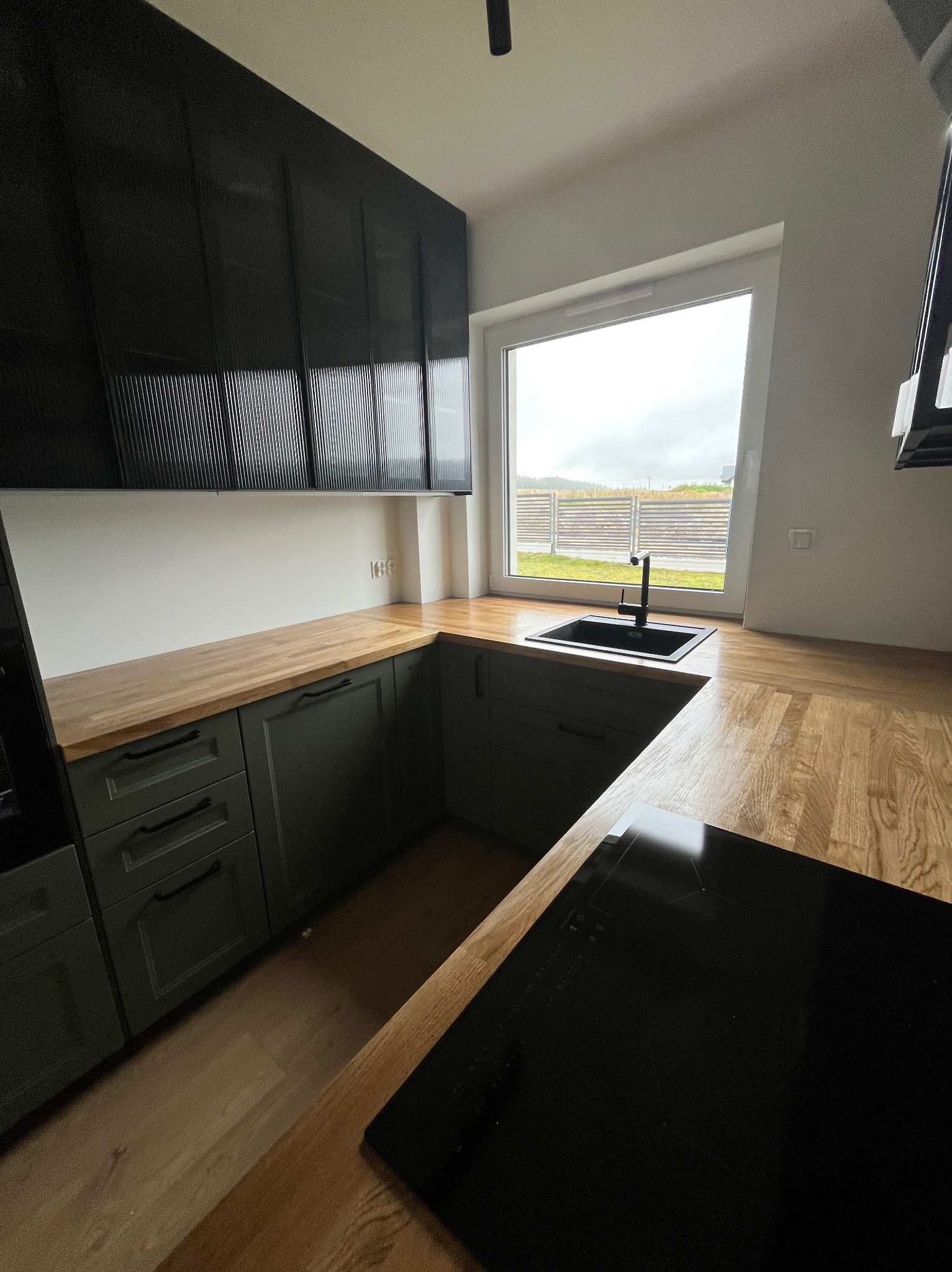 Nowoczesna kuchnia z ciemnozielonymi szafkami, drewnianym blatem i czarnym zlewem pod oknem z widokiem na zewnątrz. Minimalistyczny design, jasne wnętrze.