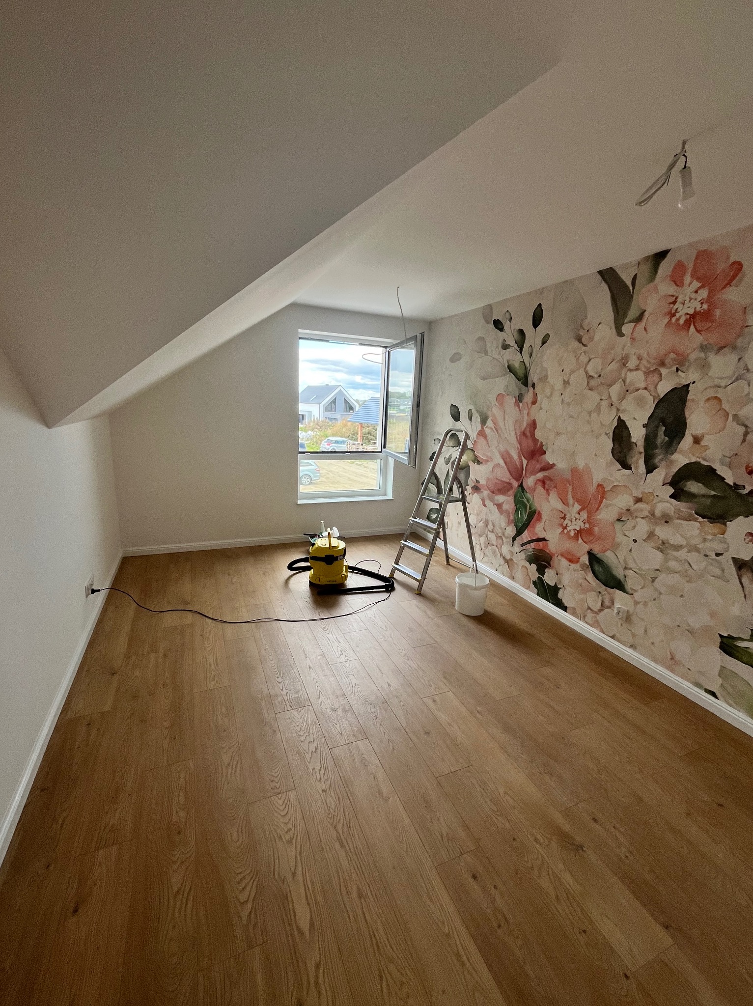 Pokój na poddaszu po remoncie: kwiecista tapeta, drewniana podłoga, drabina, odkurzacz, okno z widokiem na dom. Przygotowanie do sprzątania po remoncie.
