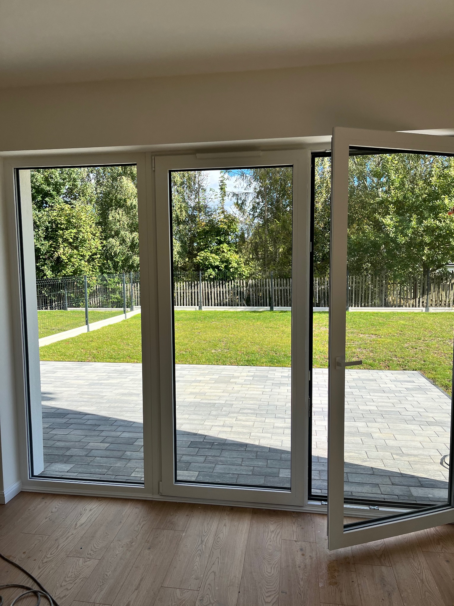 Trzyczęściowe okno tarasowe z białymi ramami, otwarte na ogród z trawnikiem i kostką brukową. Jedno skrzydło uchylone. Parkiet na podłodze wewnątrz.