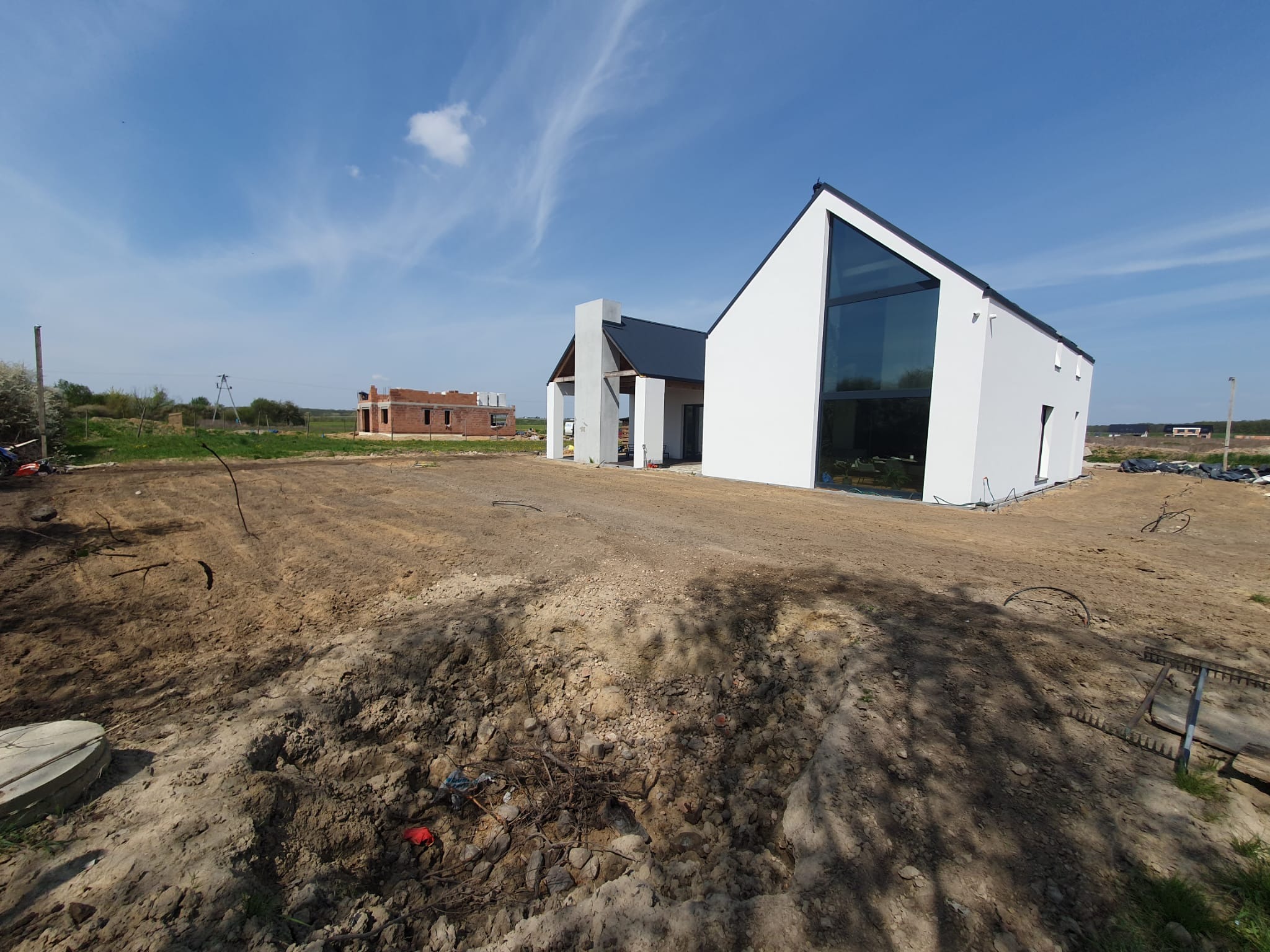 Przygotowany teren pod ogród przed nowoczesnym domem z dużym oknem. Widoczne ślady po pracach ziemnych i wyrównywaniu terenu. W tle budynek w trakcie budowy.