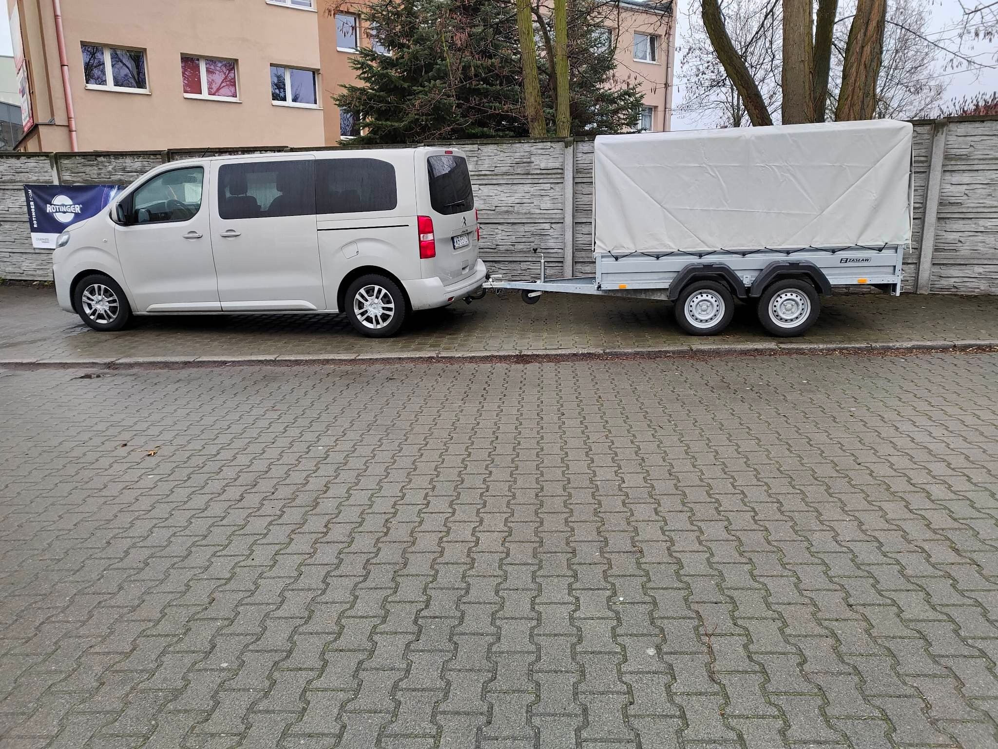 Jasny van z przyczepą z plandeką parkuje na brukowanej ulicy. Przyczepa marki Zasław. W tle budynek mieszkalny i drzewa.