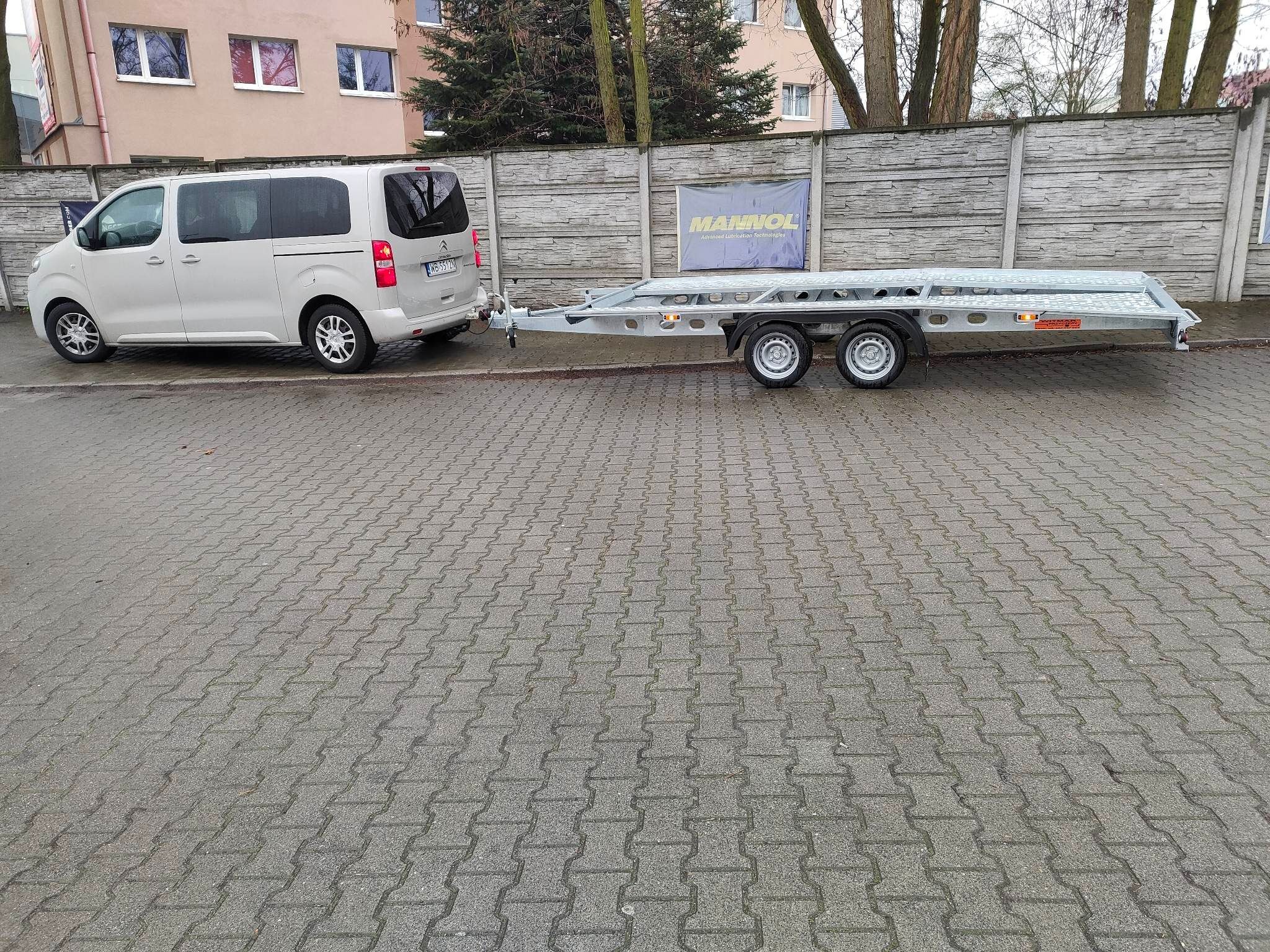 Jasnoszary van z podpiętą, dwuosiową, srebrną lawetą samochodową na kostce brukowej. W tle szary mur z reklamą Manrol.