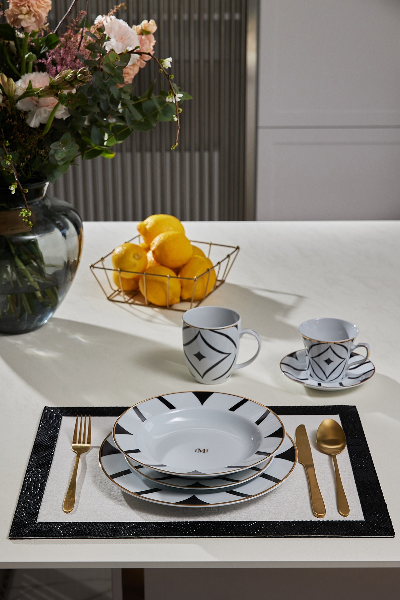 Elegancka zastawa stołowa La Mania Home w geometryczne wzory, złote sztućce, cytryny w koszyku i kwiaty w wazonie na białym stole. Minimalistyczne tło.