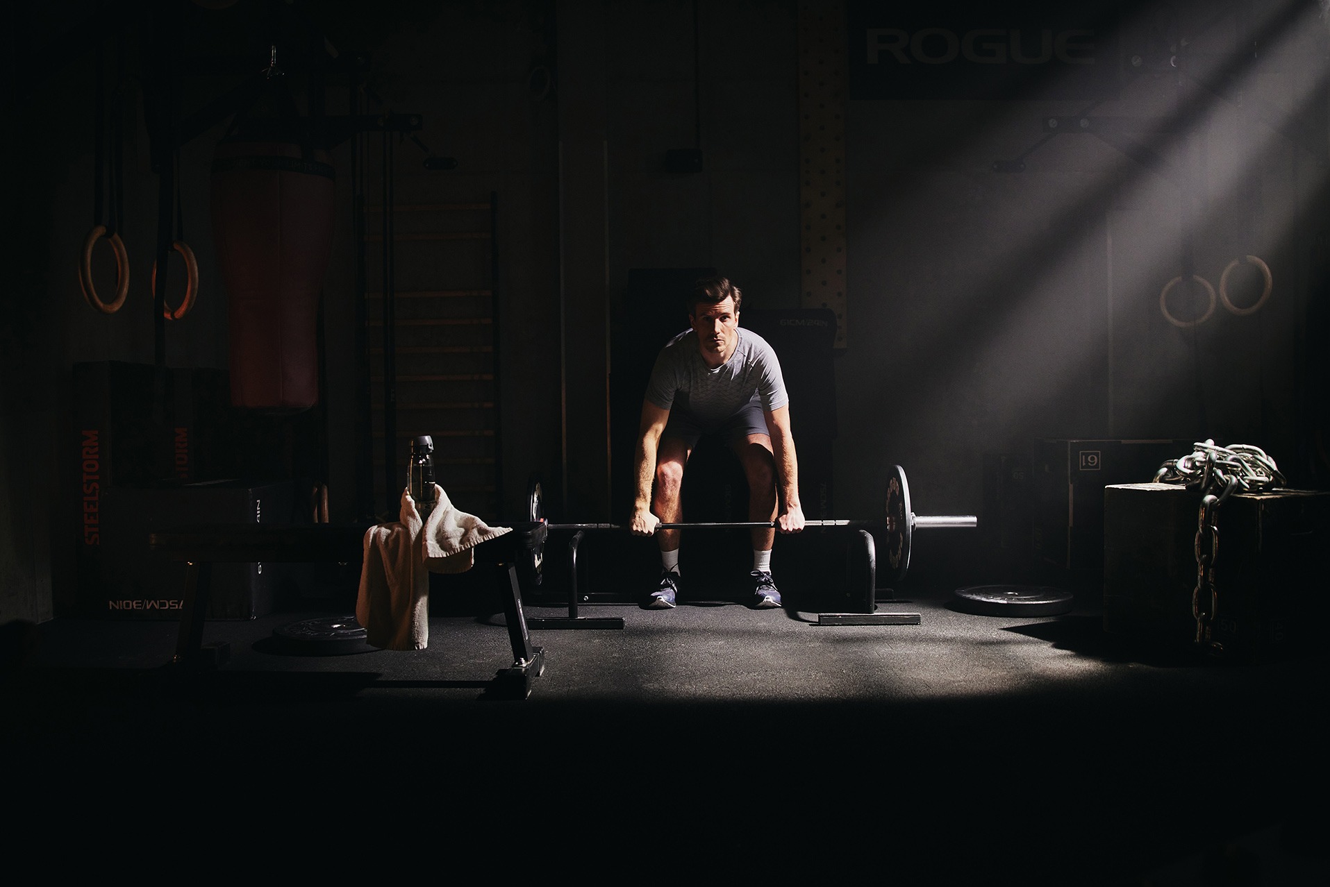 Mężczyzna przygotowuje się do podniesienia sztangi w ciemnym, profesjonalnym studio fitness z elementami wyposażenia Rogue i Steelstorm, oświetlonym punktowo.