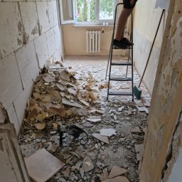 Serhii popenko - Wnętrze w trakcie wyburzania ściany z widocznym stosem gruzu, wiertarką udarową i osobą na drabinie. Fragment ściany z tapetą w tle.