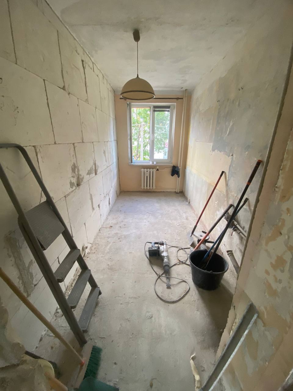 Wnętrze pokoju w trakcie prac. Widoczne ślady po usuniętych tapetach, ściana z bloczków, drabina, sprzęt budowlany i okno z widokiem na zieleń.