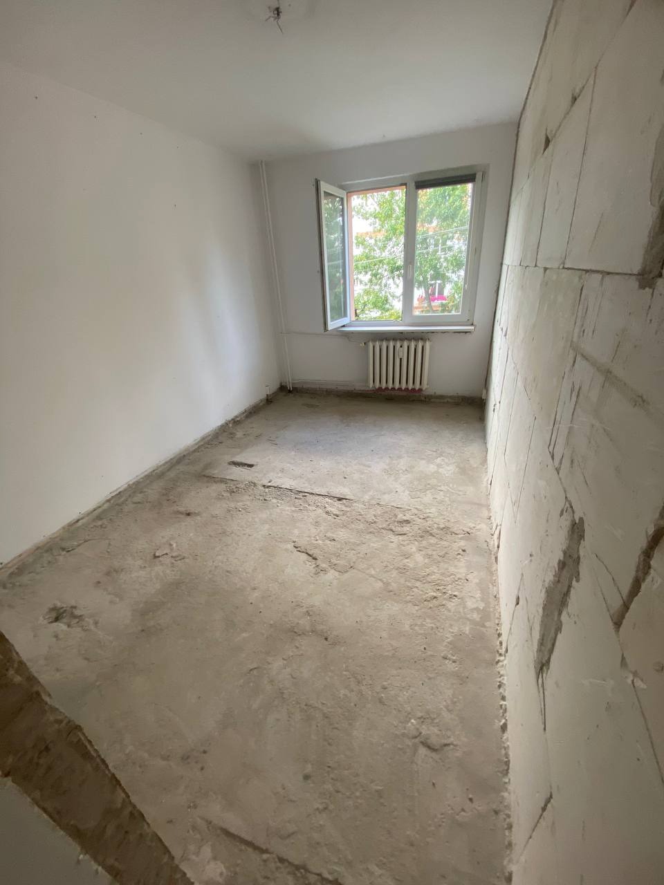 Pokój po usunięciu warstw podłogi i części ściany, widoczny gruz i odsłonięta ściana z pustaków. Okno z widokiem na zieleń. Grzejnik pod oknem.