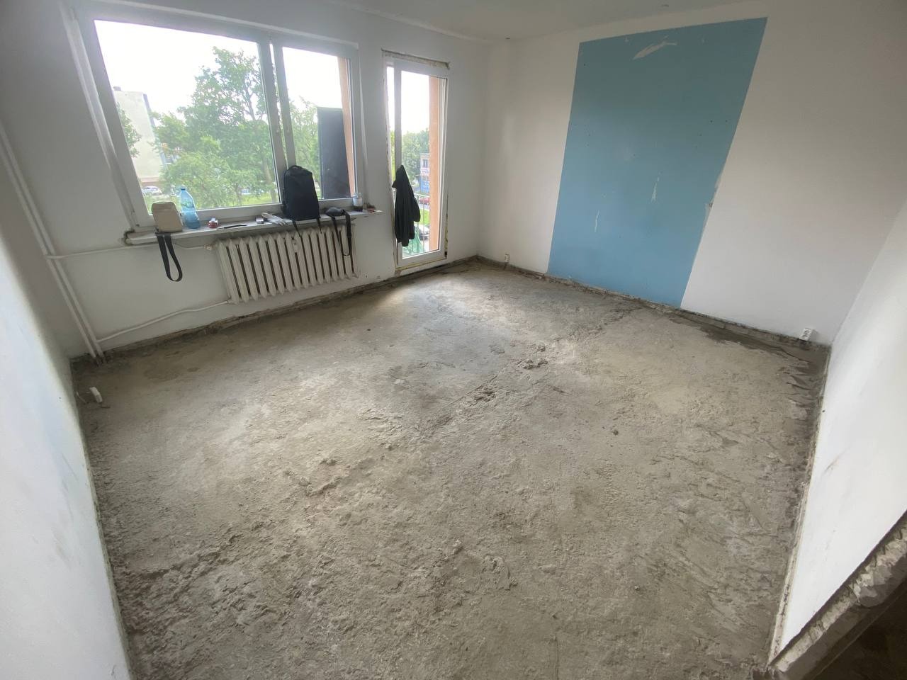 Pokój po demontażu podłogi, widoczne ślady po kleju i nierówności. Ściany z fragmentami farby i tynku. Grzejnik i okno z widokiem na zieleń.