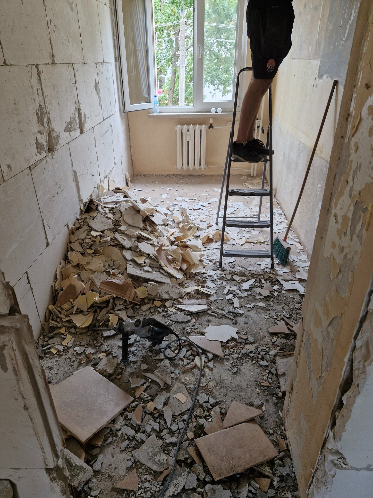 Wnętrze w trakcie wyburzania ściany z widocznym stosem gruzu, wiertarką udarową i osobą na drabinie. Fragment ściany z tapetą w tle.