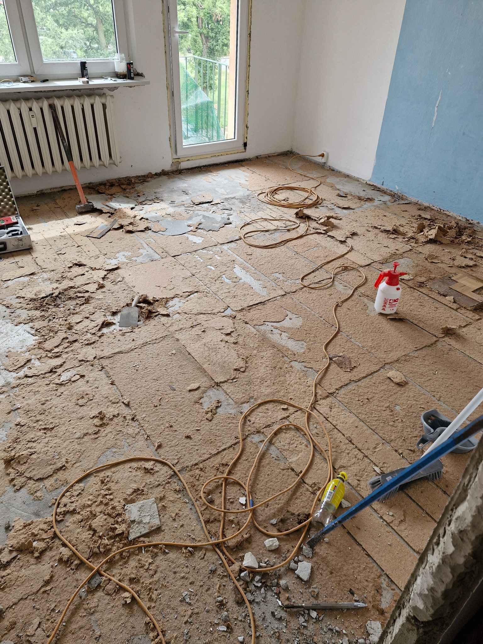 Pokój w trakcie usuwania starej podłogi: widoczne fragmenty paneli, narzędzia (młotek, szpachelka) oraz przewody na podłodze. Ściany częściowo pomalowane na niebiesko.