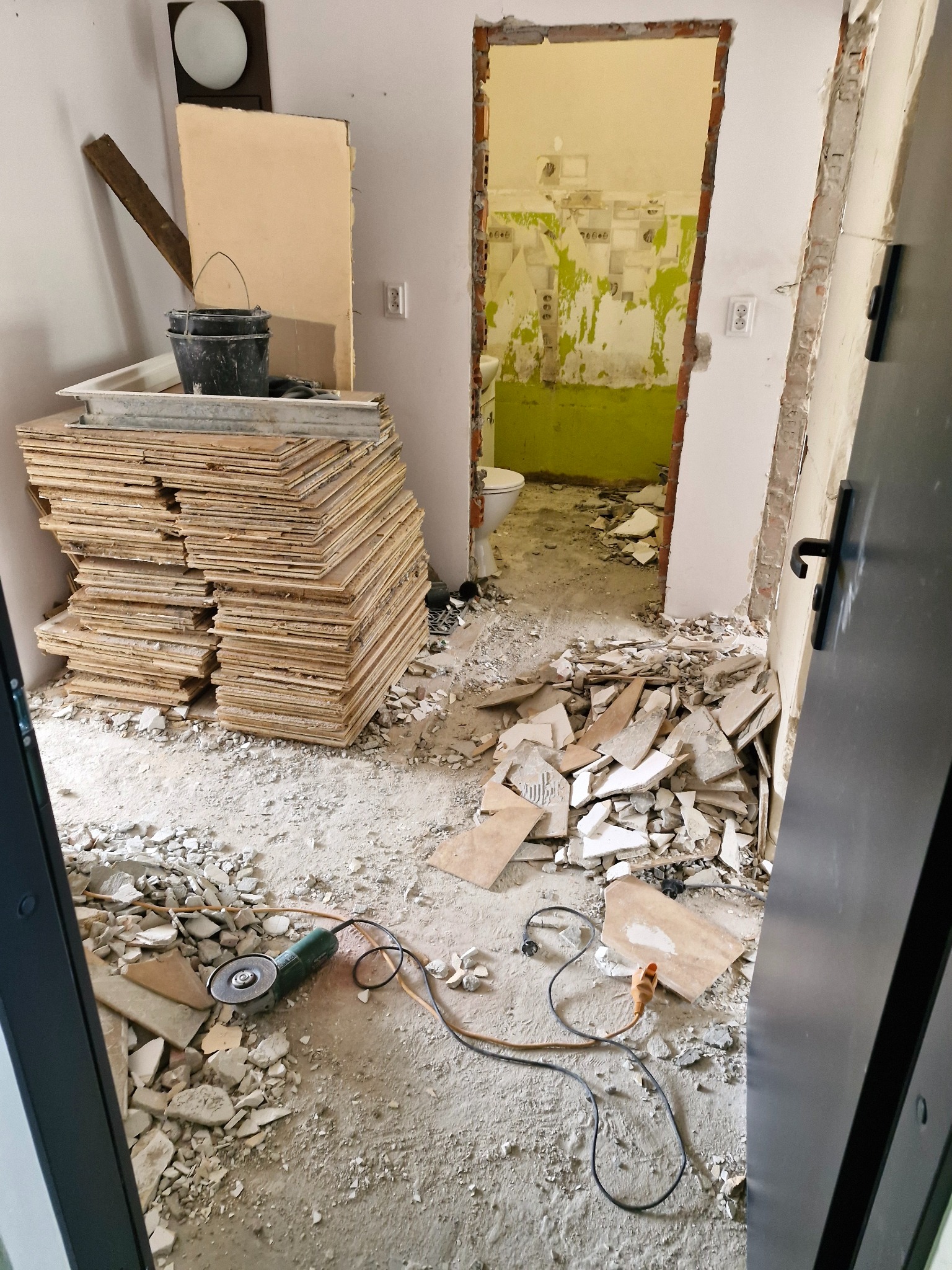 Po demontażu: sterta płyt, gruz i szlifierka kątowa w pomieszczeniu z widoczną, odartą ścianą w tle. Otwarta przestrzeń po wyburzeniu.