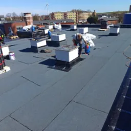 Pracownik w trakcie uszczelniania komina na płaskim dachu pokrytym papą termozgrzewalną. Widoczne narzędzia, butla gazowa i budynki w tle.