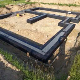 Izolacja fundamentów z papy i styroduru na piaskowym podłożu, z widocznymi prętami zbrojeniowymi i żółtym pojemnikiem. Ujęcie z góry, słoneczny dzień.