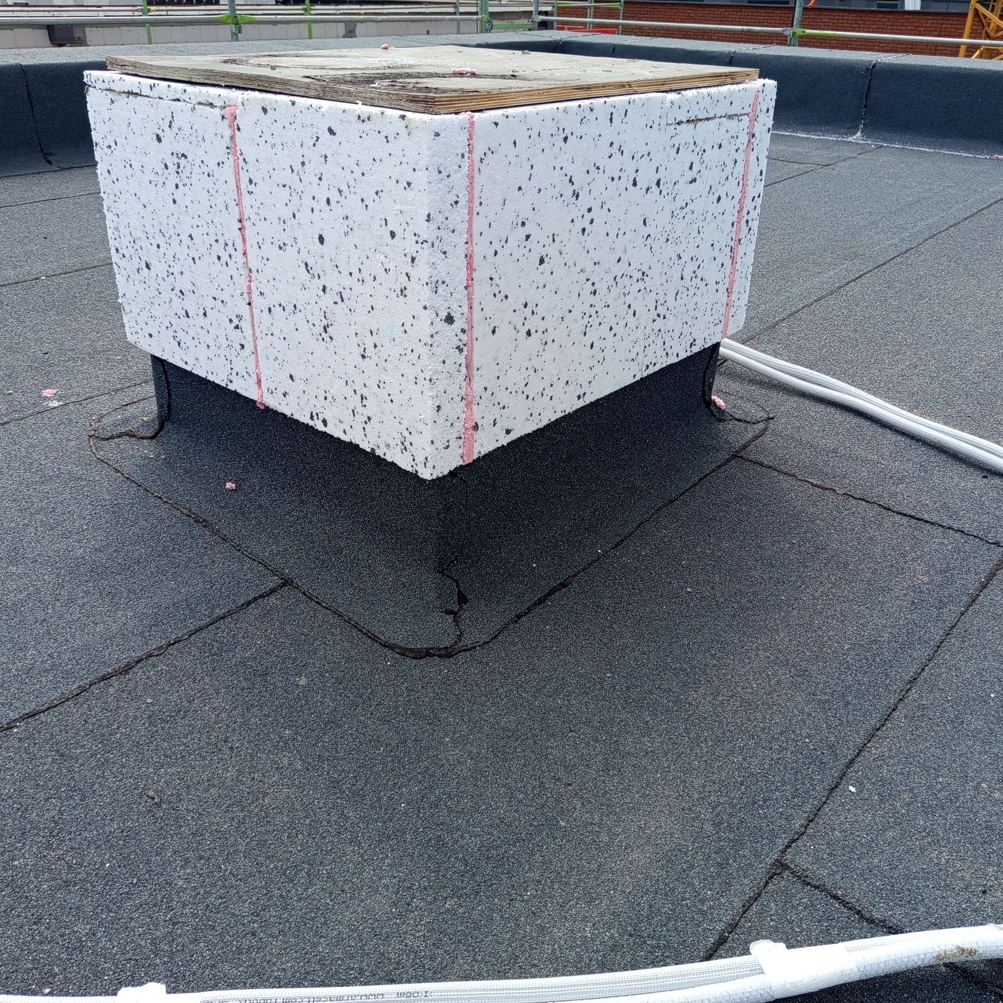 Biała obudowa z izolacją na dachu pokrytym papą. Widoczne uszczelnienia i fragment instalacji. Kąt ujęcia z góry.