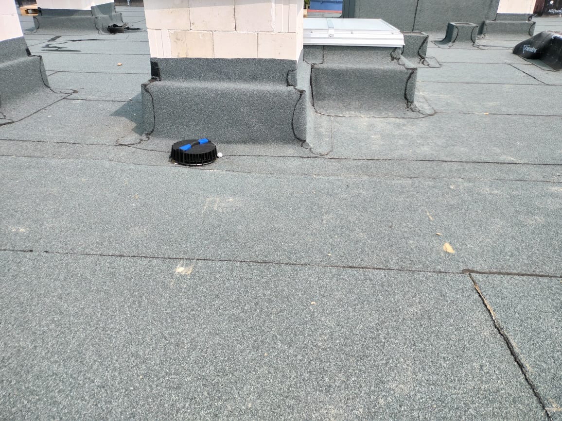 Szary dach pokryty papą z kominami i elementami wentylacyjnymi. Widoczne łączenia papy i detale wykończeniowe. Dach płaski z elementami wystającymi.