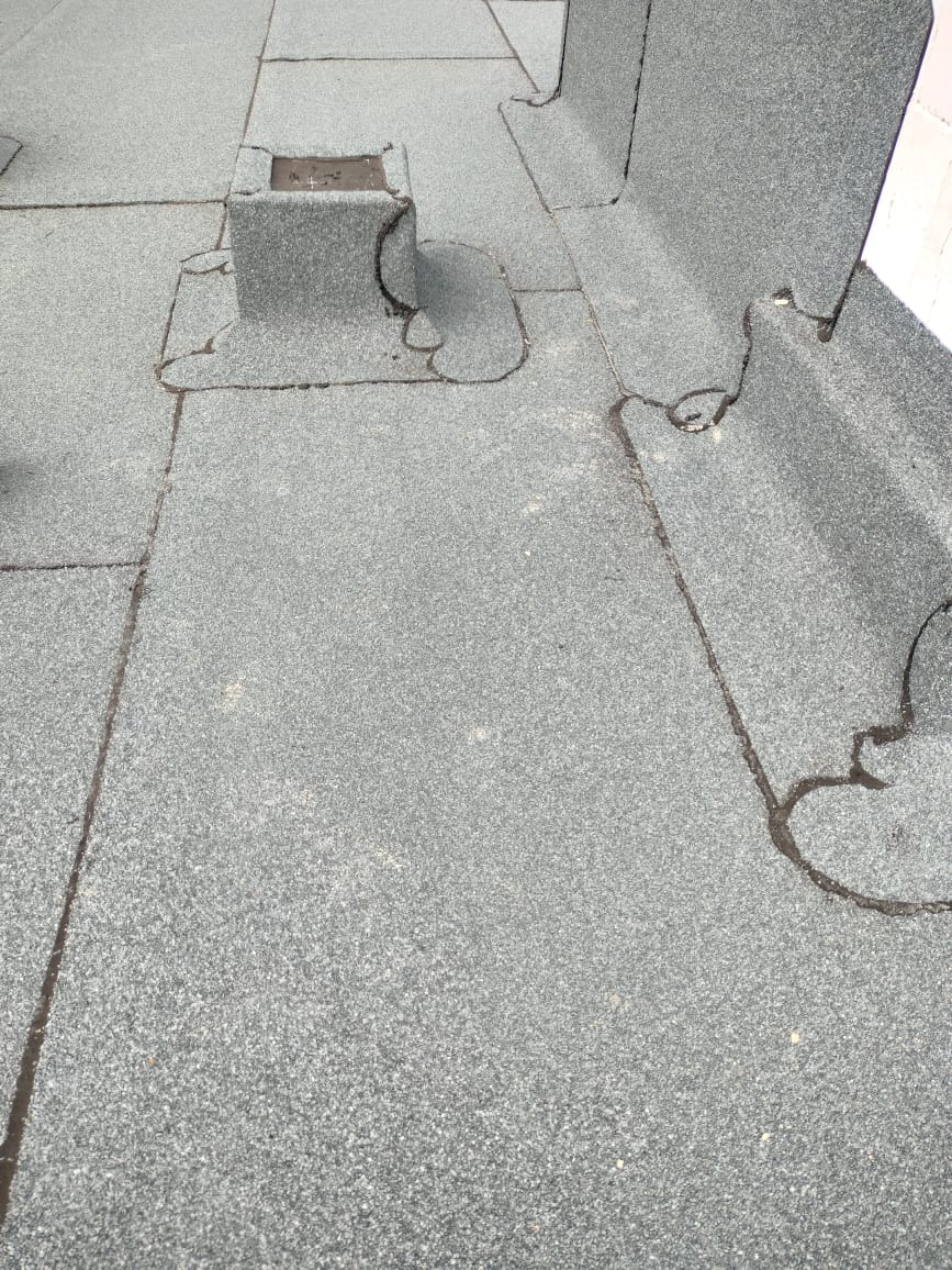 Szczegółowe ujęcie płaskiego dachu pokrytego papą z widocznymi obróbkami wokół komina i krawędzi. Szara kolorystyka i drobna faktura materiału.